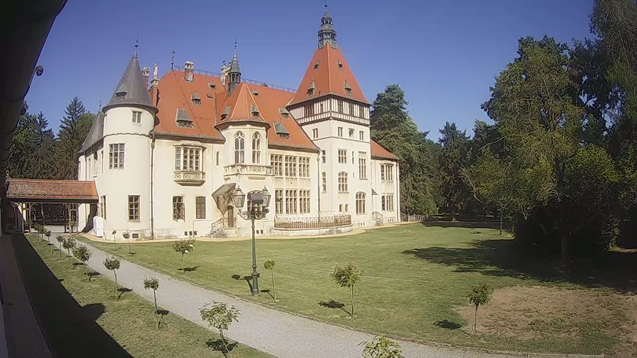 A large, light-colored castle with a red-tiled roof stands on a grassy lawn under a clear blue sky.