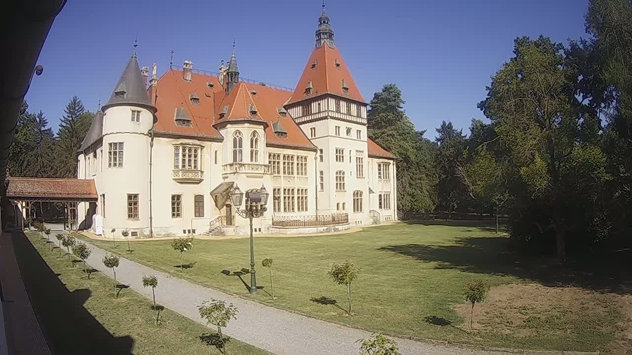 A large, light-colored castle with a red roof and multiple towers stands on a grassy lawn under a clear blue sky, next to a paved pathway and surrounded by trees.