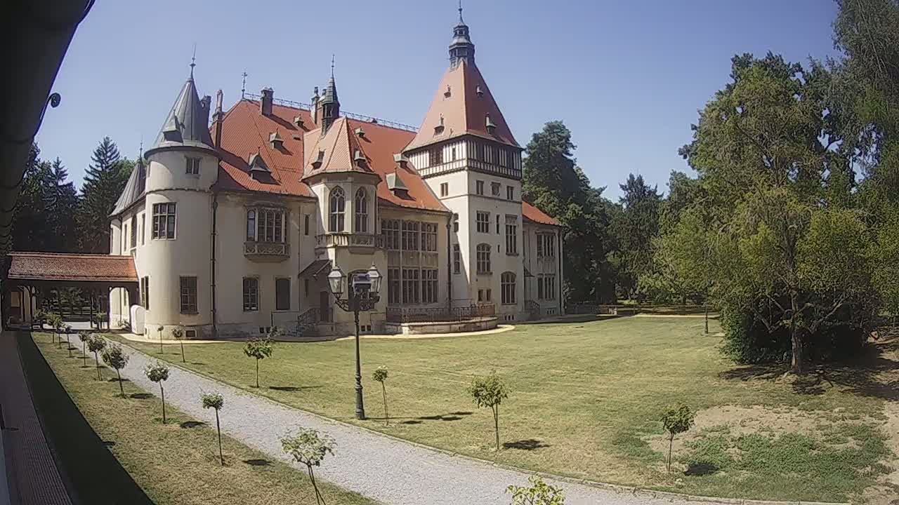 A large, light-colored castle with a red roof and multiple towers stands on a grassy lawn under a clear blue sky, next to a paved pathway and surrounded by trees.