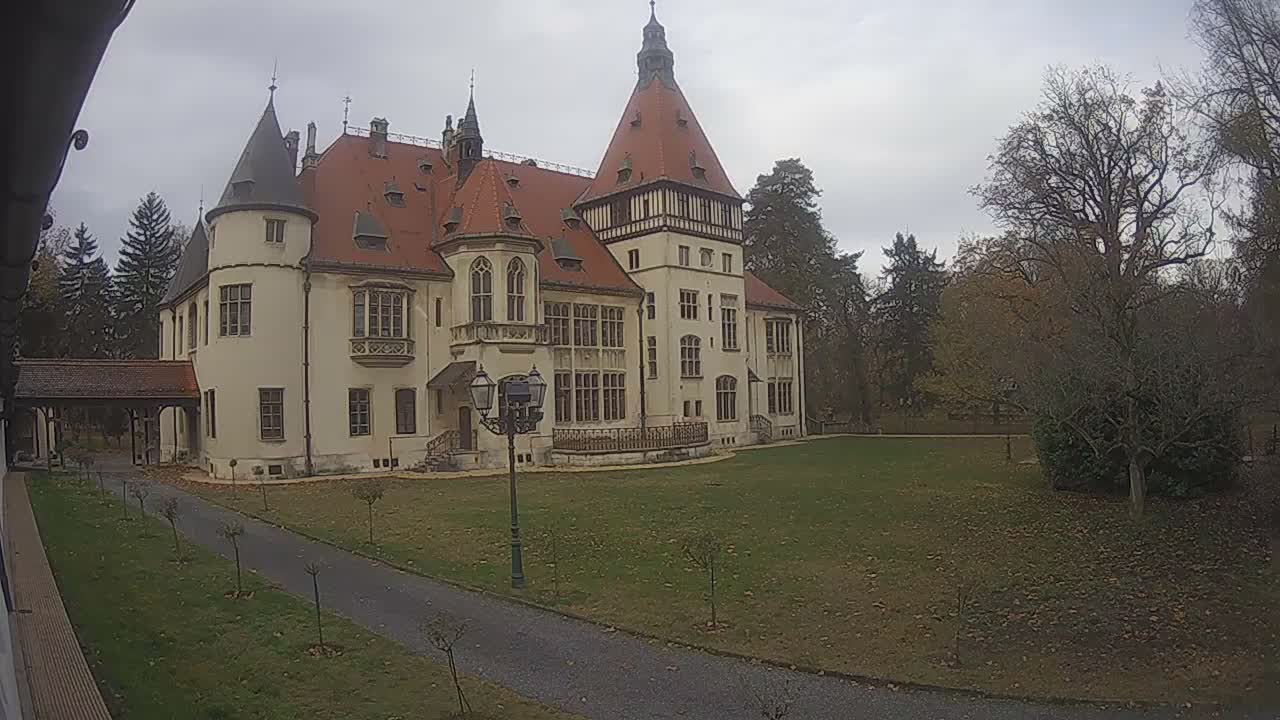 A large, light-colored castle with a red roof and multiple towers stands on a grassy lawn under a clear blue sky, next to a paved pathway and surrounded by trees.