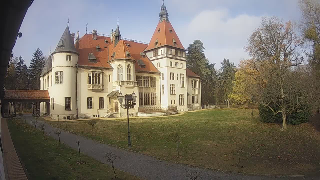 An elegant, cream-colored castle-like building with prominent reddish-orange roofs and multiple turrets is set amidst a spacious lawn and autumn trees under a partly cloudy sky.
