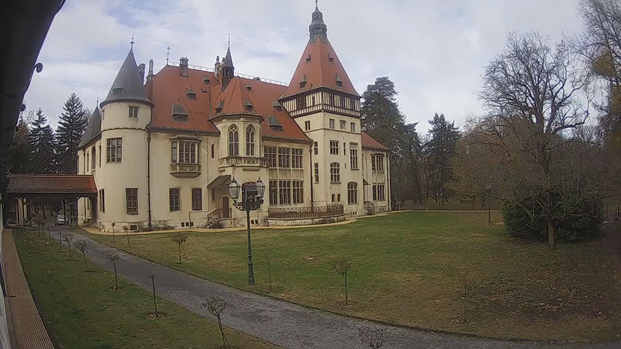 A grand, cream-colored castle featuring red-tiled roofs and multiple spires stands amidst a spacious green lawn with a gravel path, surrounded by a mix of bare deciduous and evergreen trees, all under a cloudy sky.