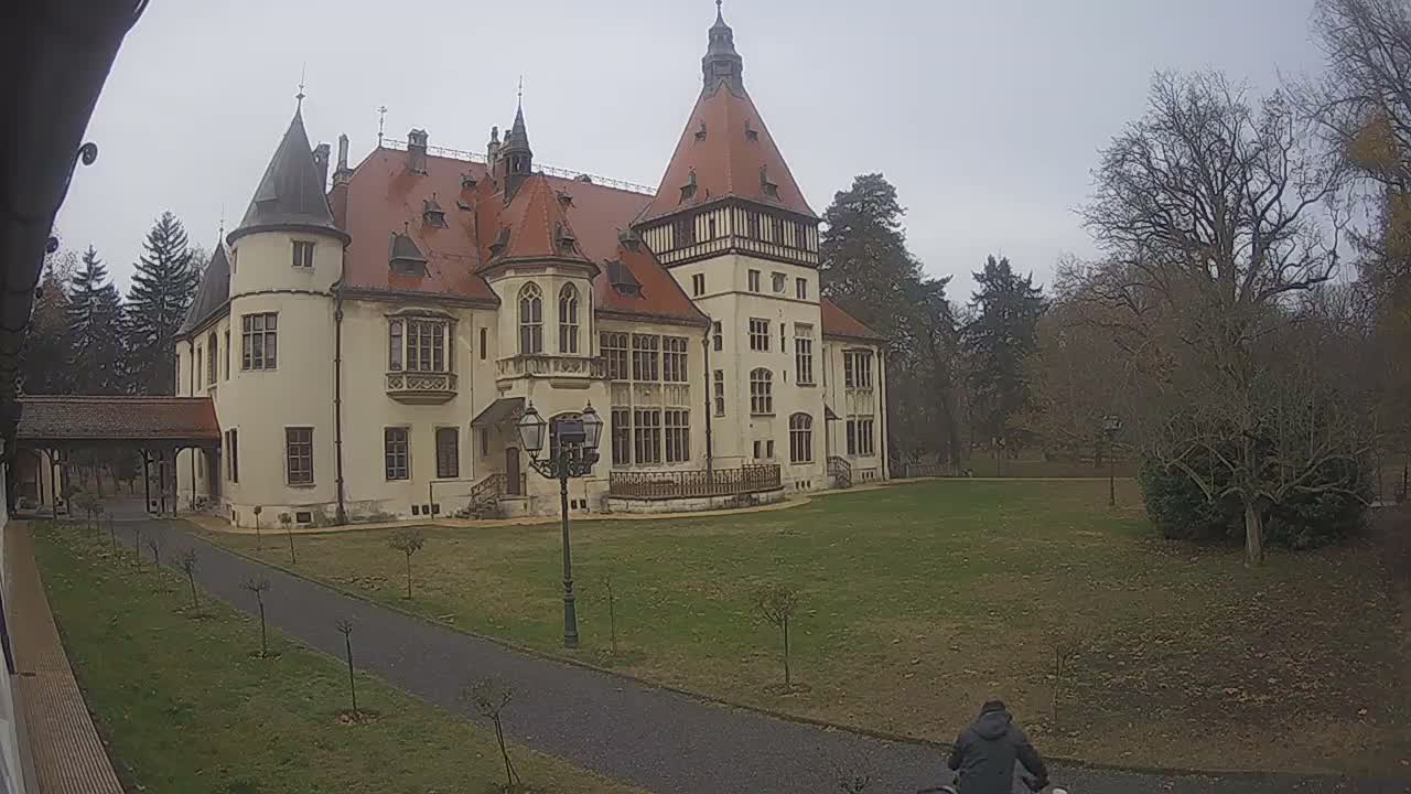 A person rides a bicycle down a gravel path past a grand, cream-colored manor with red roofs and multiple towers, surrounded by a grassy lawn and bare trees under an overcast sky.