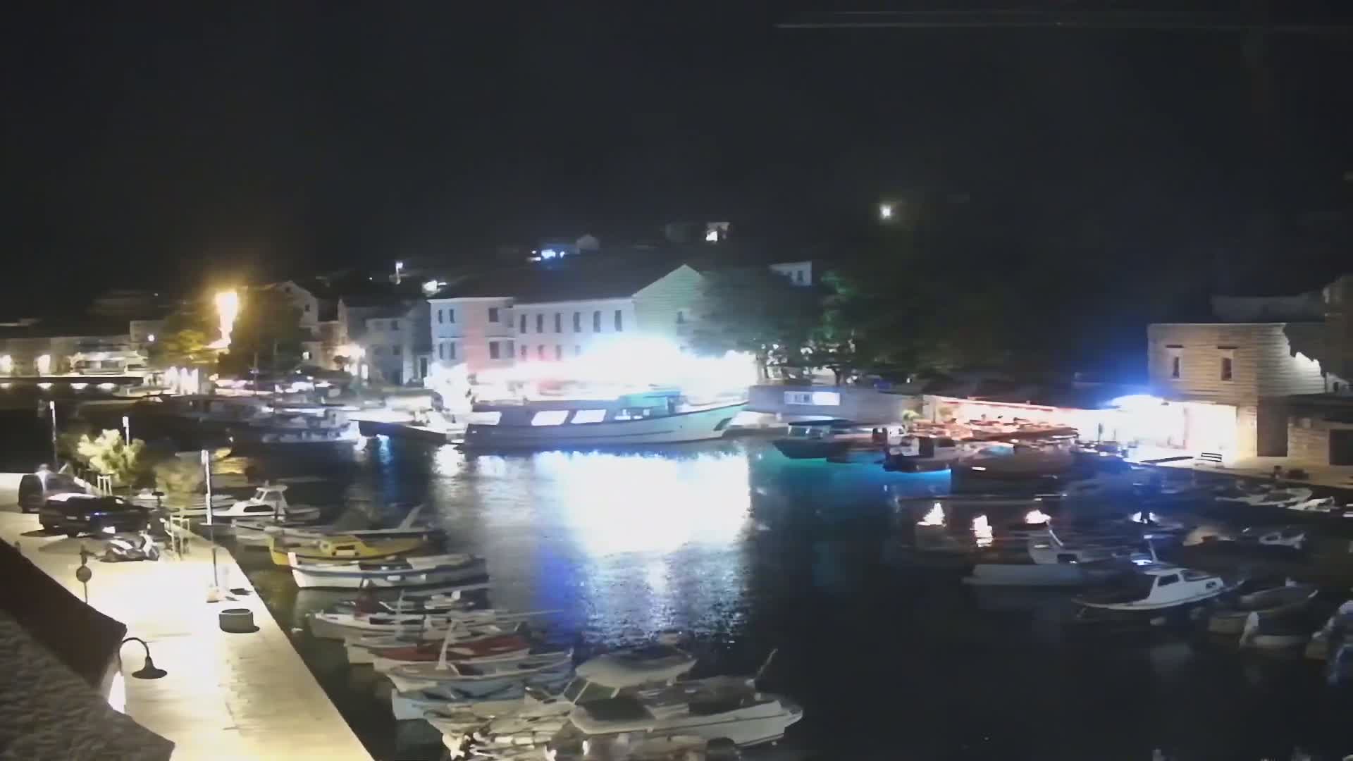A nighttime harbor scene shows numerous boats docked along a waterfront lined with buildings, under a clear, dark sky.