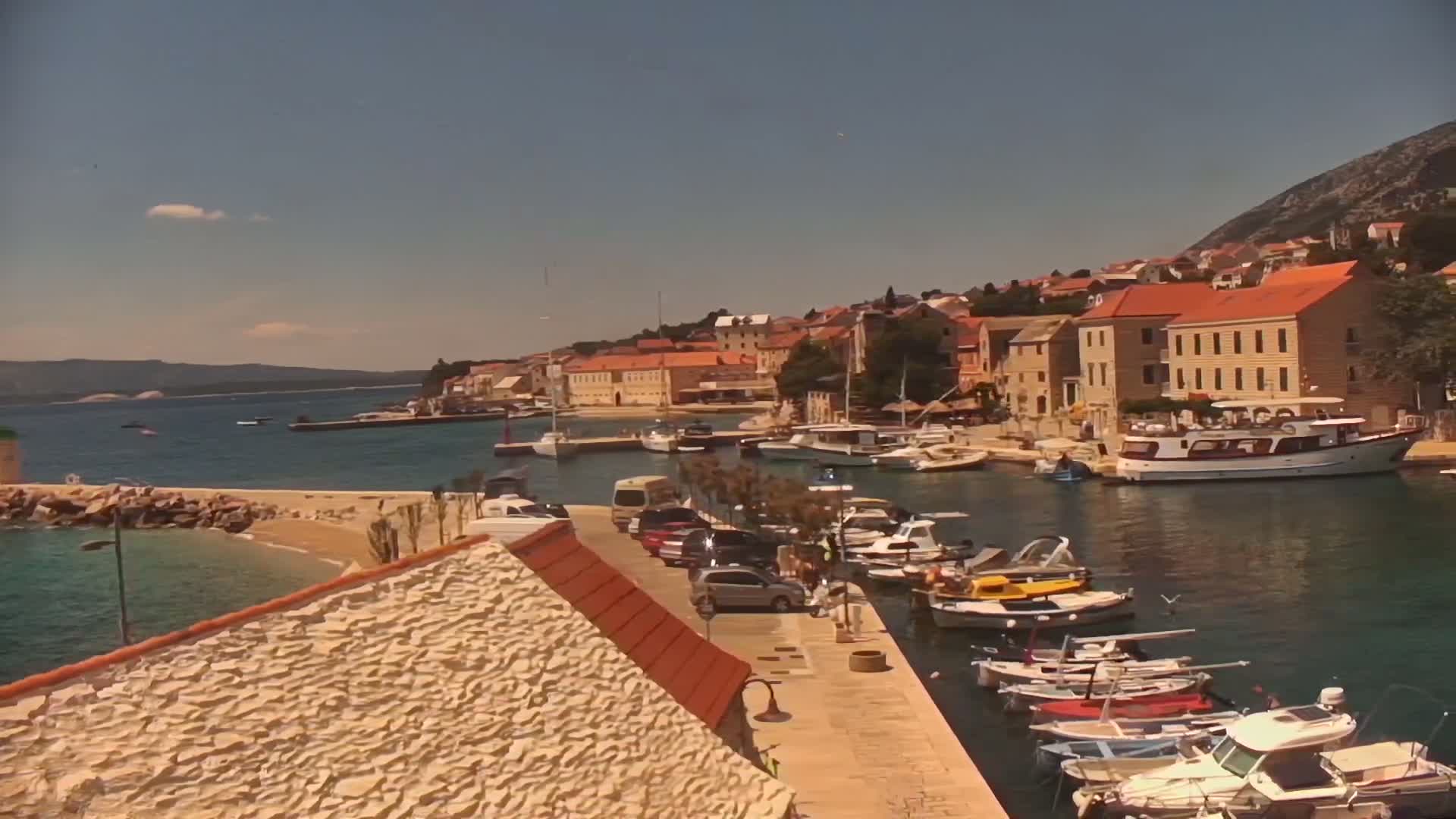 A sunny day reveals a harbor town nestled against a hillside, with numerous boats docked and cars parked along a stone walkway.