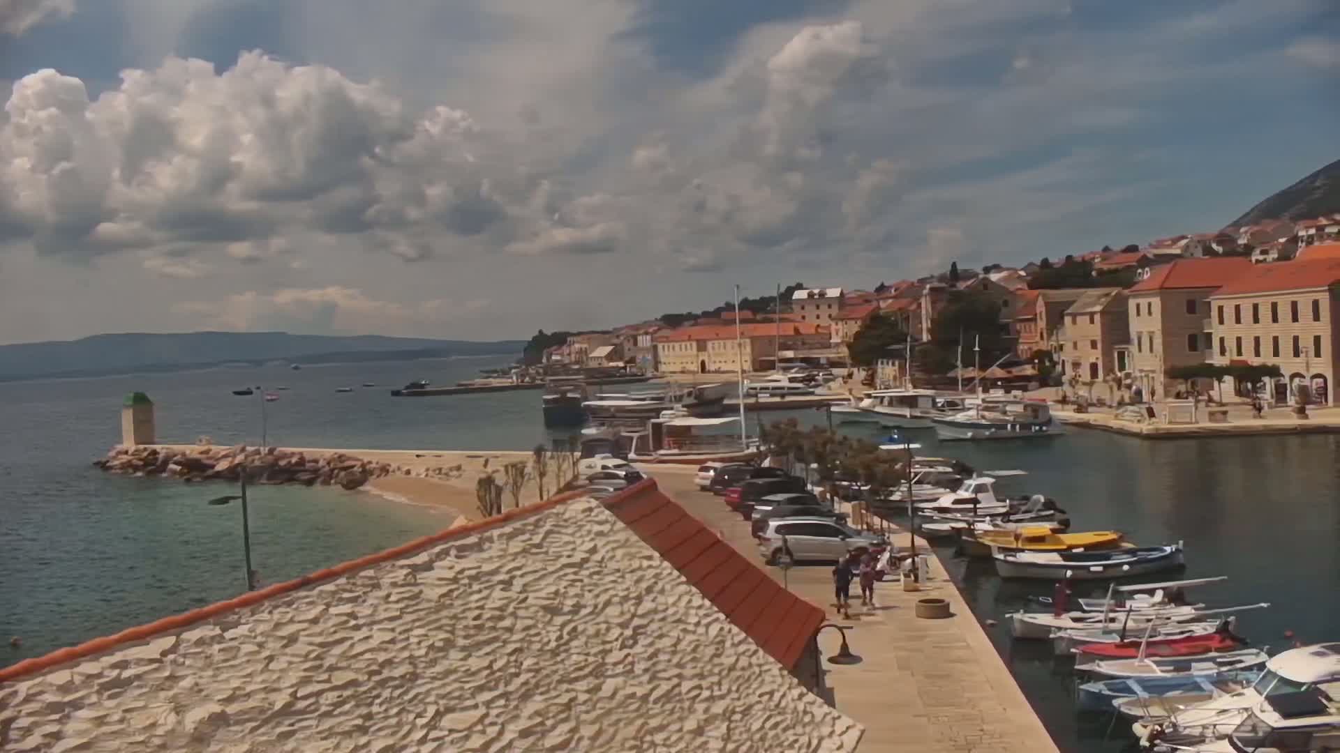A partly sunny day reveals a picturesque harbor town with numerous boats docked along a calm waterway,  adjacent to a stone building and a small sandy beach.