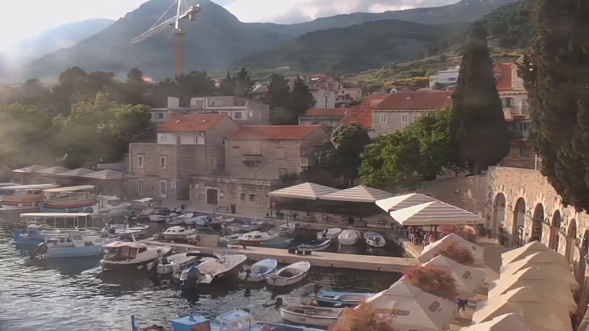 A harbor filled with numerous boats is nestled amongst stone buildings at the base of a mountain range under a partly cloudy sky.