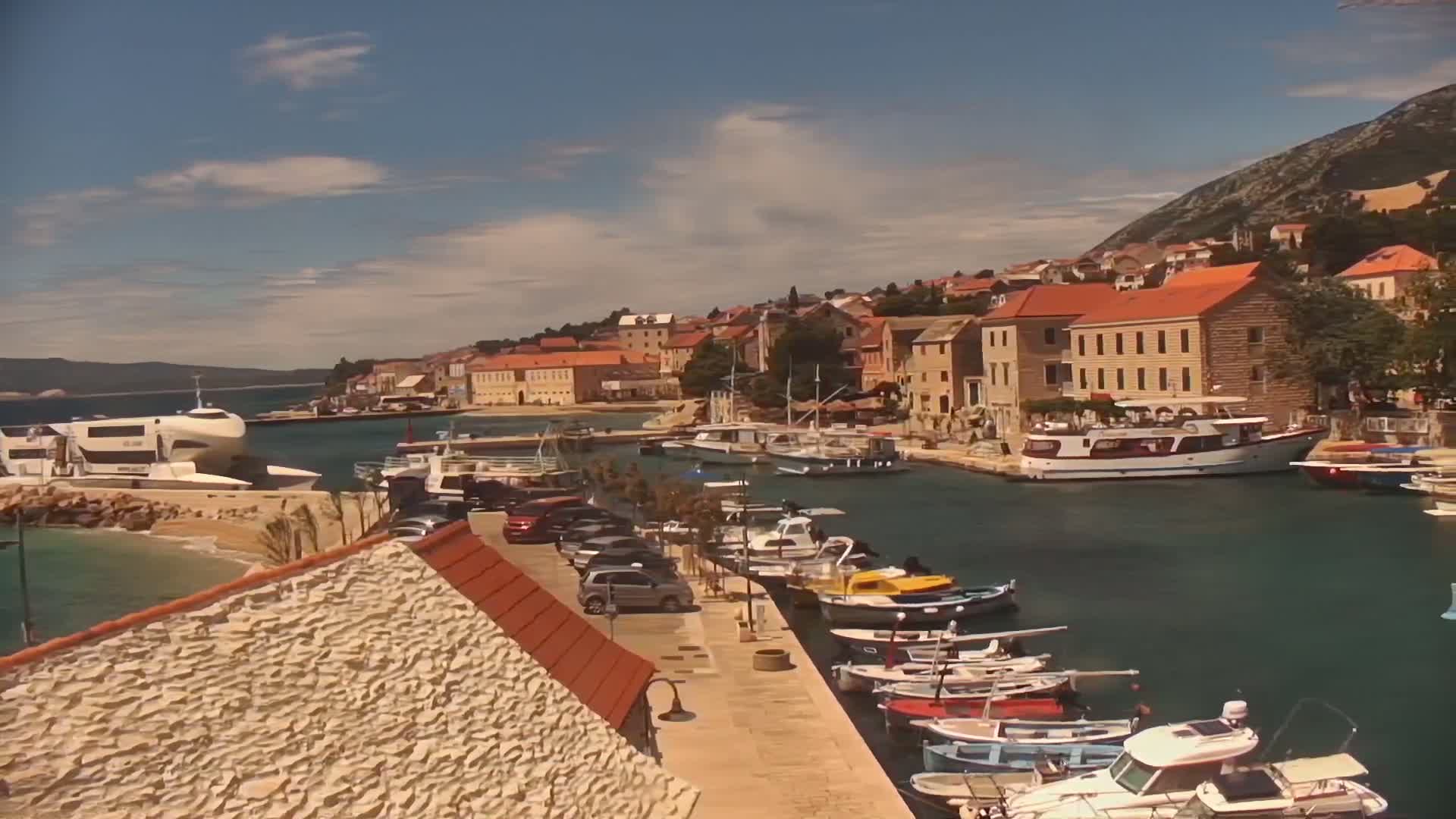 A harbor filled with numerous boats and yachts is nestled alongside a town of terracotta-roofed buildings under a partly sunny sky.