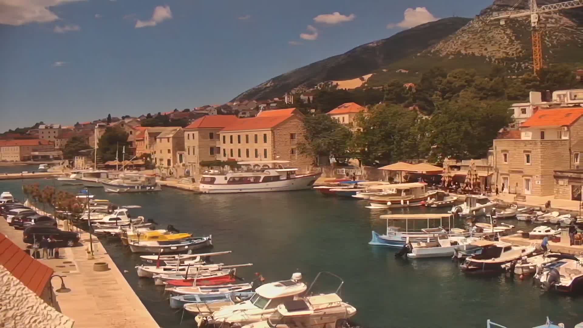 A harbor filled with numerous boats of various sizes is nestled against a waterfront town, backed by a mountain under a partly sunny sky.