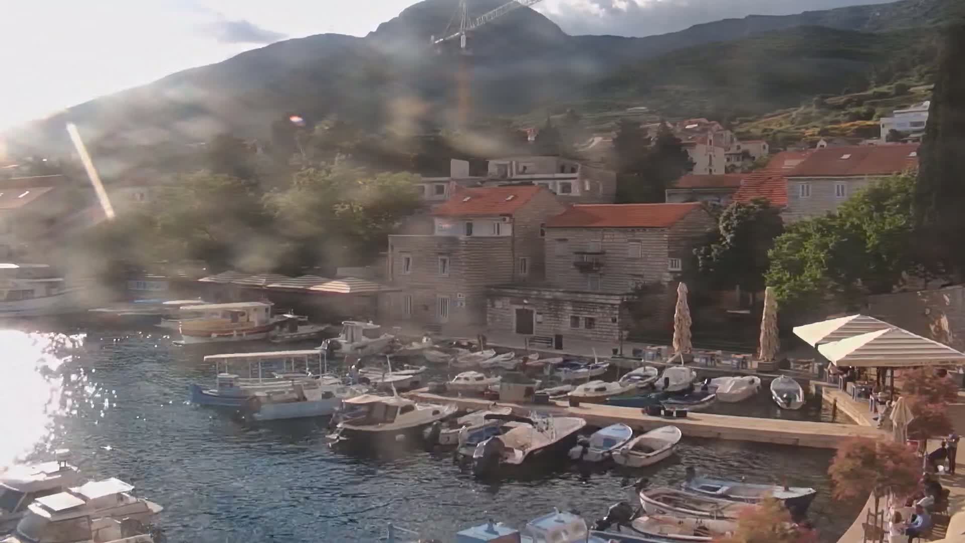 A harbor filled with numerous boats is nestled beside a town situated at the foot of a mountain, under a partly sunny sky with hazy conditions.
