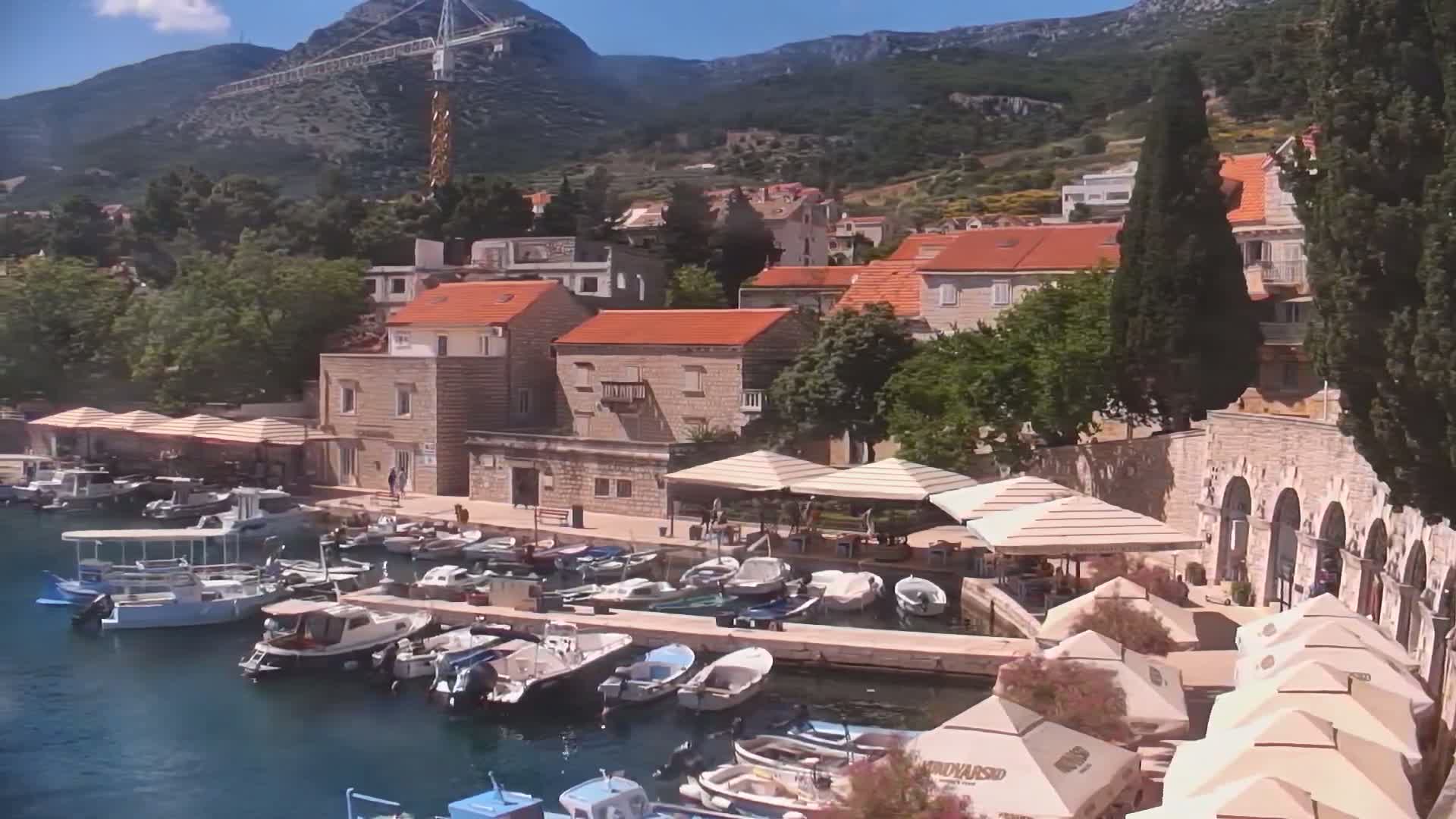 A sunny harbor scene shows numerous small boats docked along a stone quay, with buildings and outdoor seating areas lining the waterfront, all nestled beneath mountains.