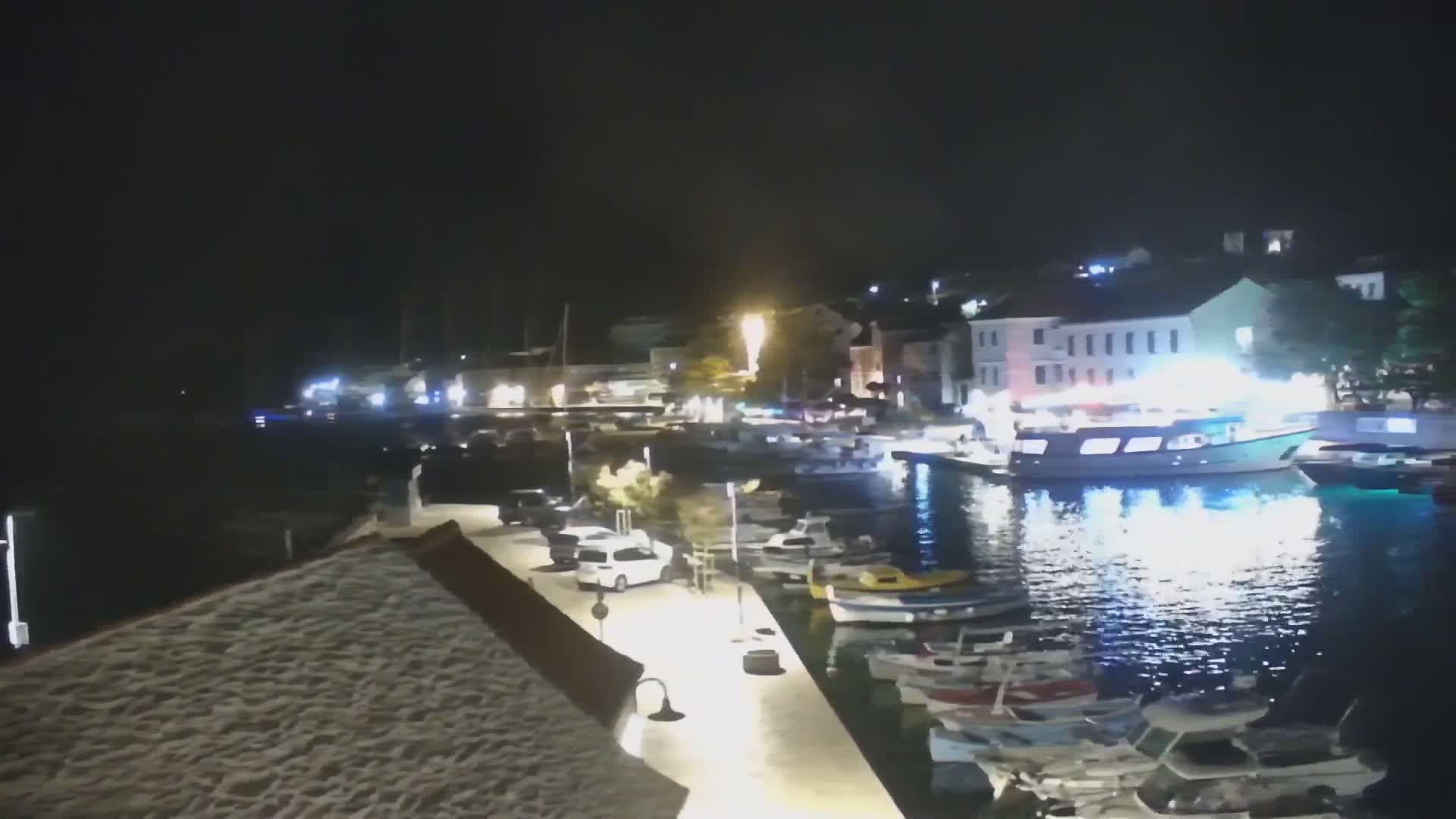 A nighttime view of a harbor filled with boats, cars parked along a waterfront walkway, and buildings illuminated along the shoreline.