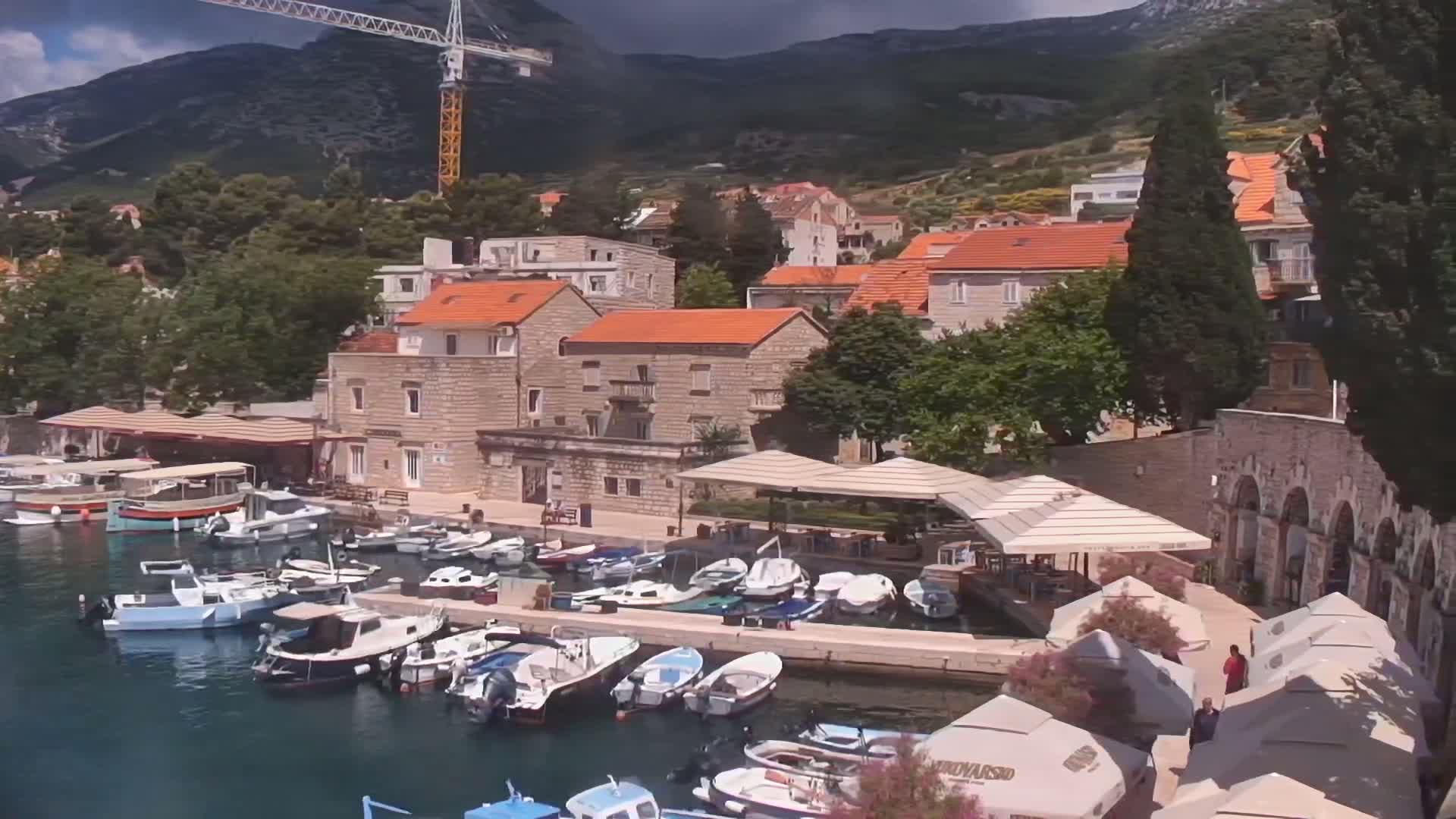 A harbor filled with numerous small boats is nestled beside a stone village at the foot of a mountain under partly cloudy skies.