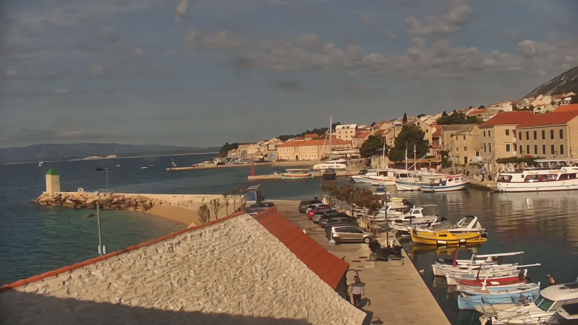 A harbor filled with boats is nestled next to a town of light-colored buildings under a partly cloudy sky.