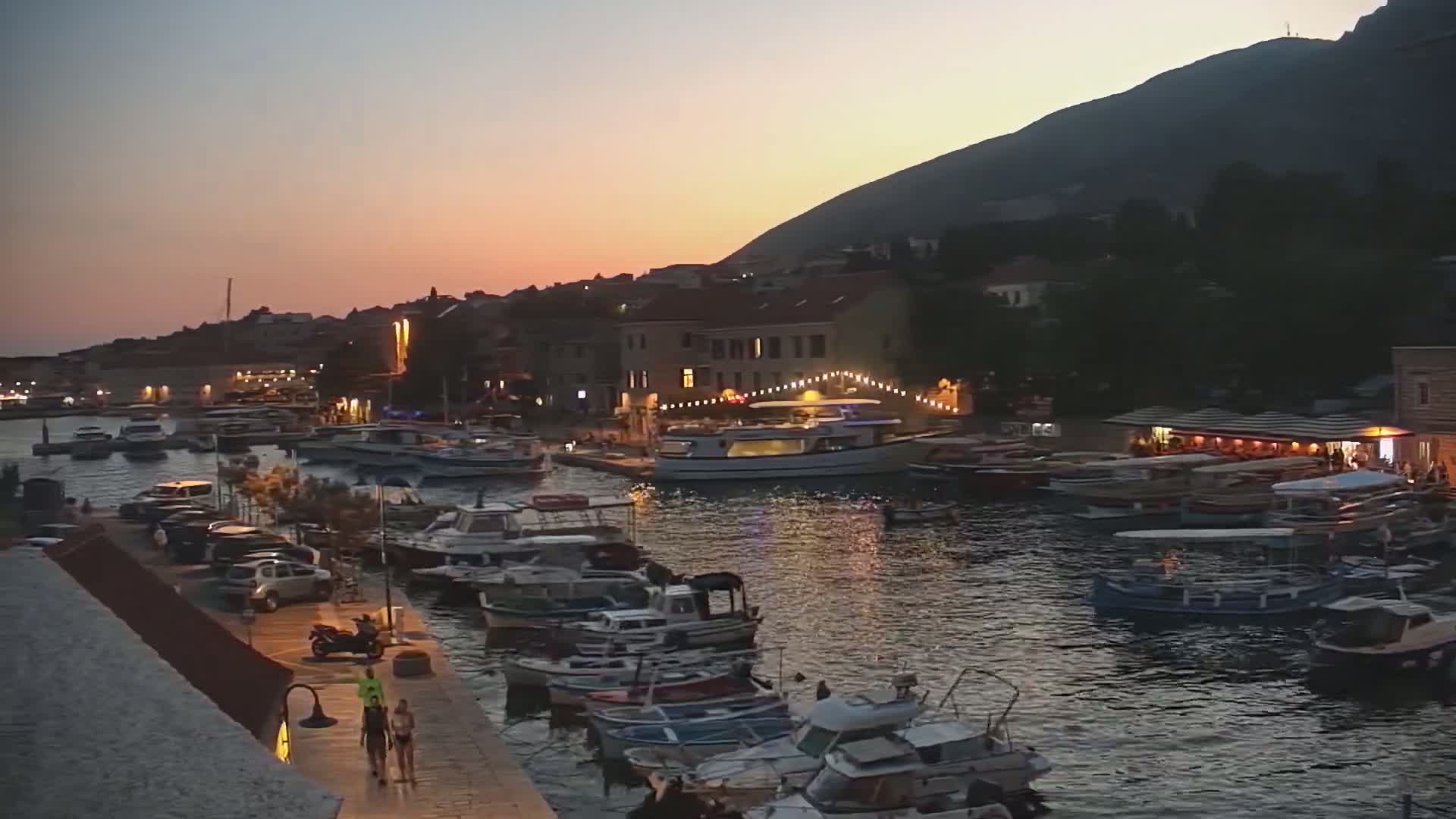 A harbor filled with numerous boats of various sizes, under a cloudy sky, is nestled beside a town of stone buildings against a mountain backdrop.