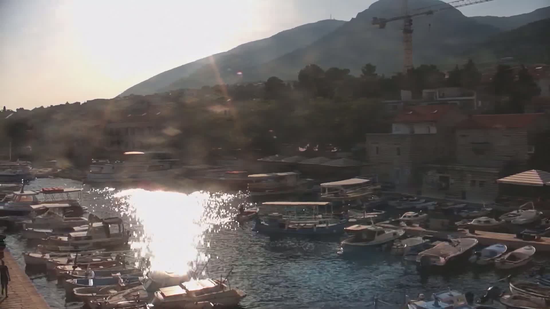 A harbor filled with numerous boats is nestled beneath a sun-drenched mountain, with a construction crane visible in the background.