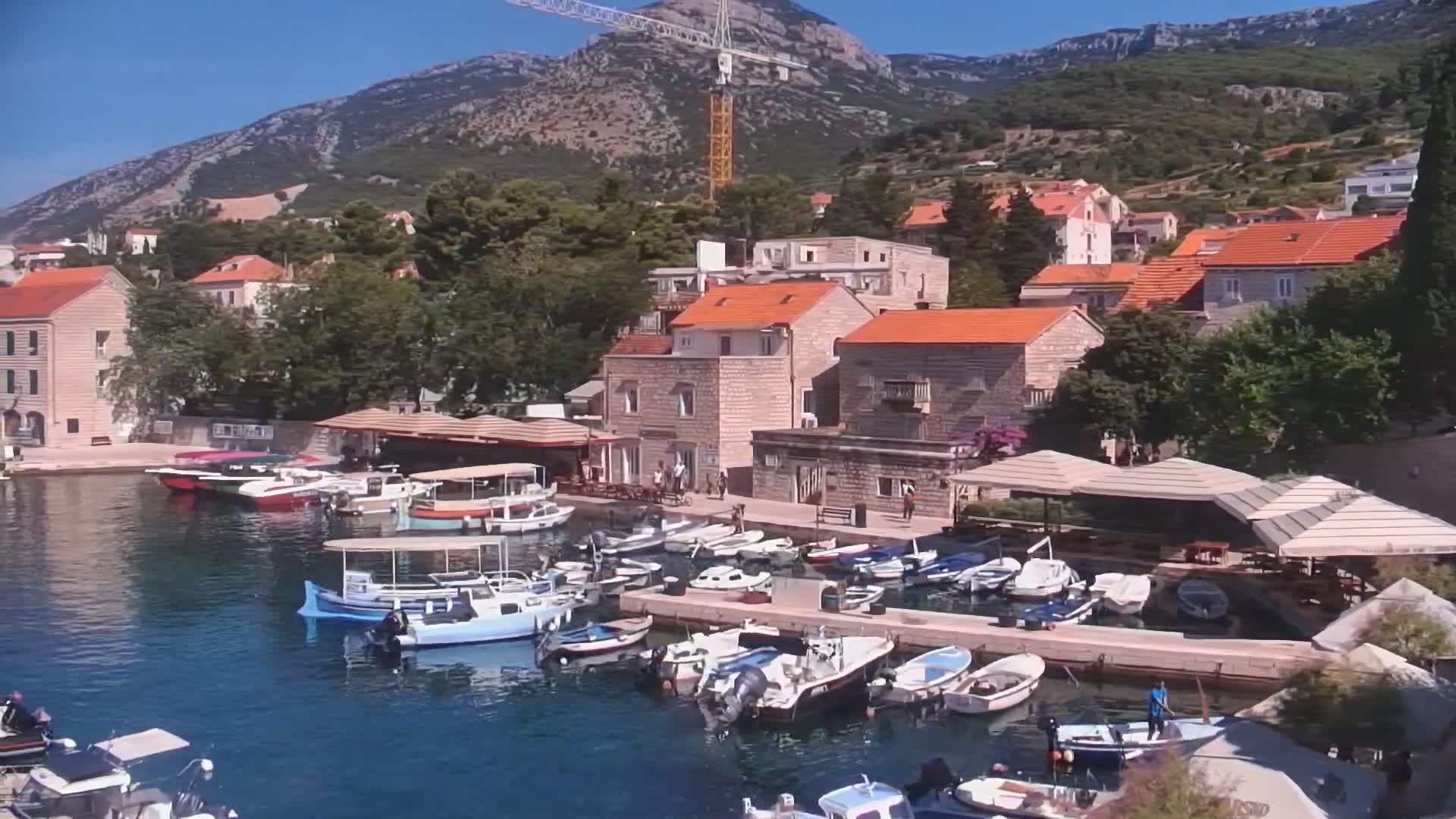 A harbor filled with numerous boats is nestled amongst a village of stone buildings with red-tiled roofs, all situated against a backdrop of mountains under a clear, sunny sky.