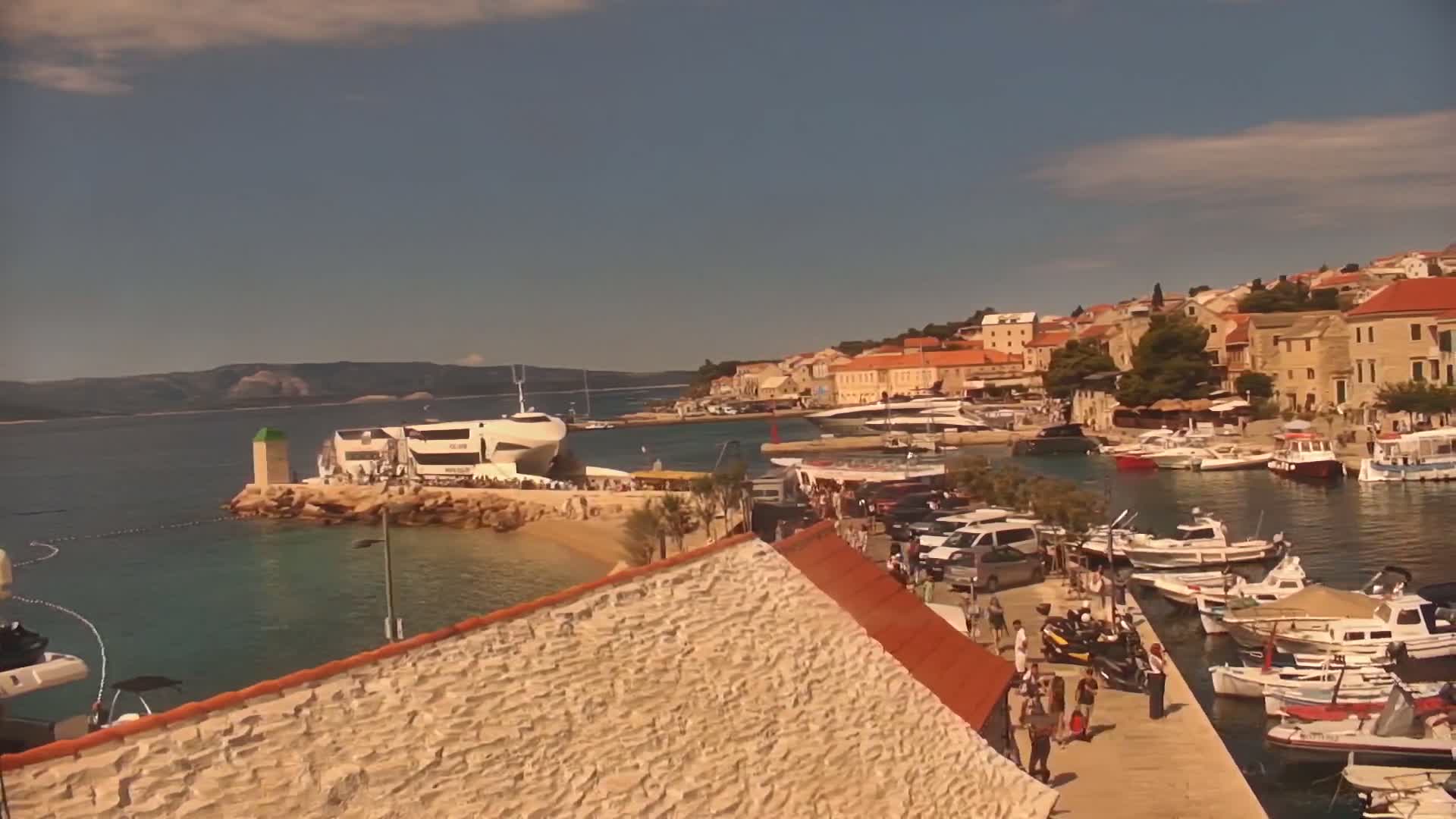 A sunny day shows a harbor filled with boats and a small town of stone buildings under a partly cloudy sky, with a large white yacht docked near a small beach.