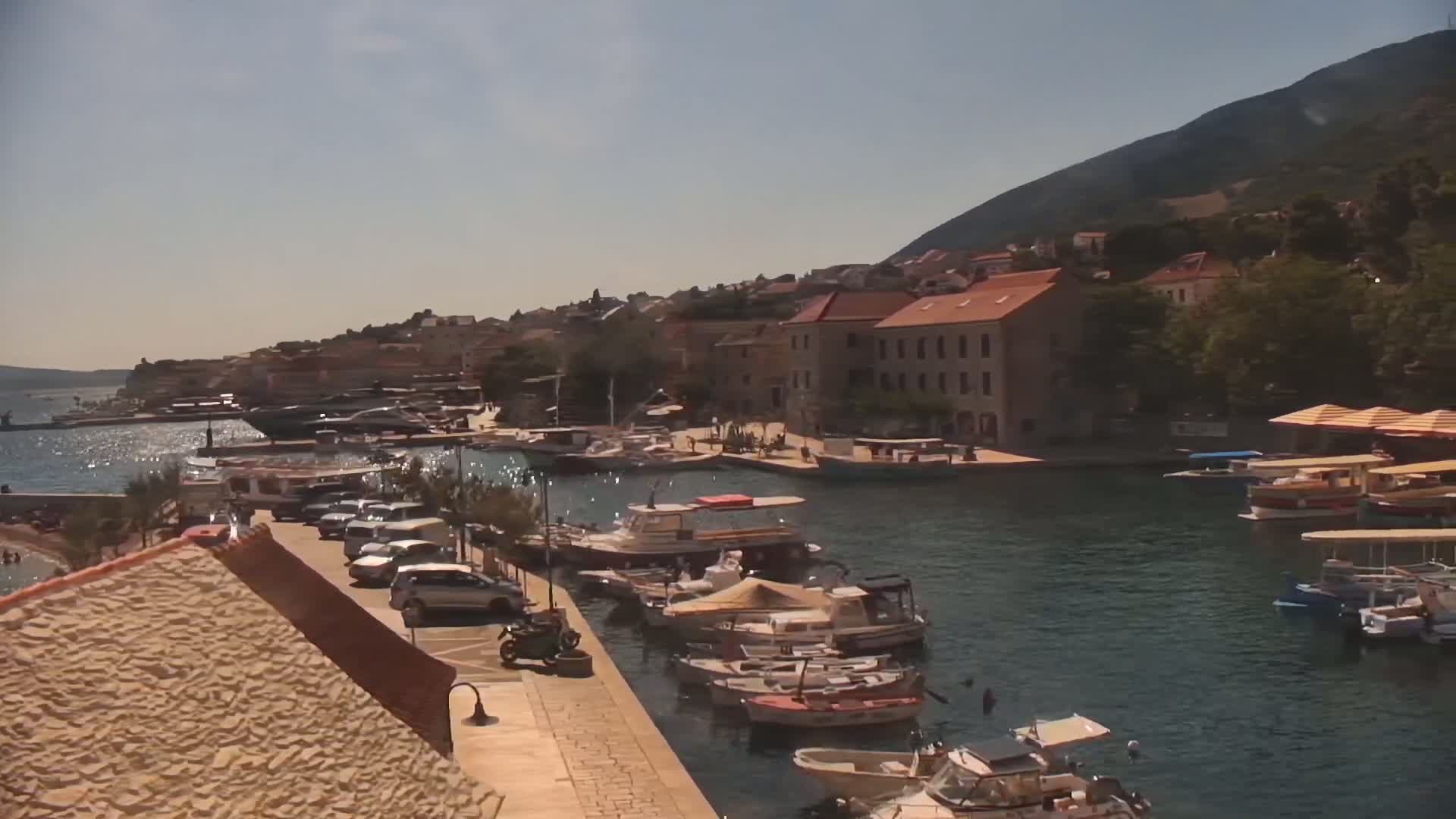 A sunny day shows a harbor filled with boats and a small town of stone buildings under a partly cloudy sky, with a large white yacht docked near a small beach.