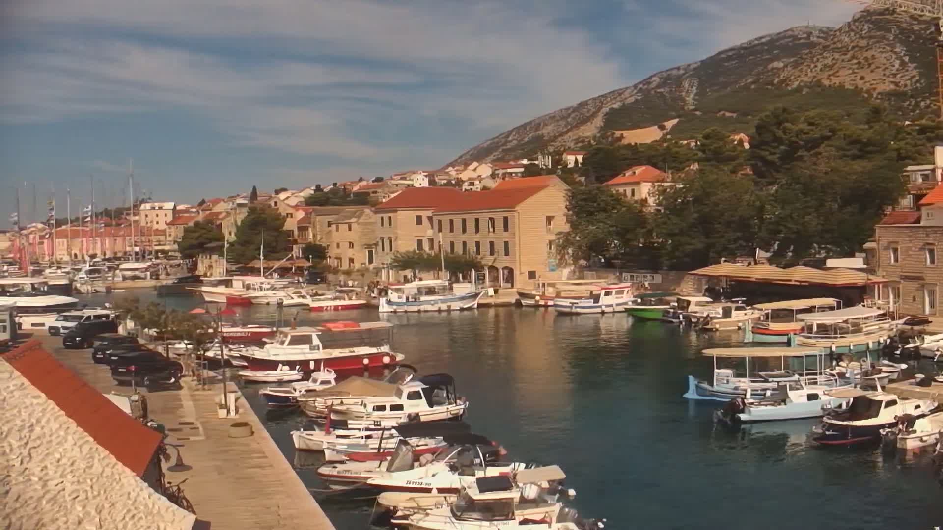 A harbor filled with numerous boats and yachts is nestled beside a town of low, tan buildings, all situated beneath a clear, sunny sky and a mountainous backdrop.