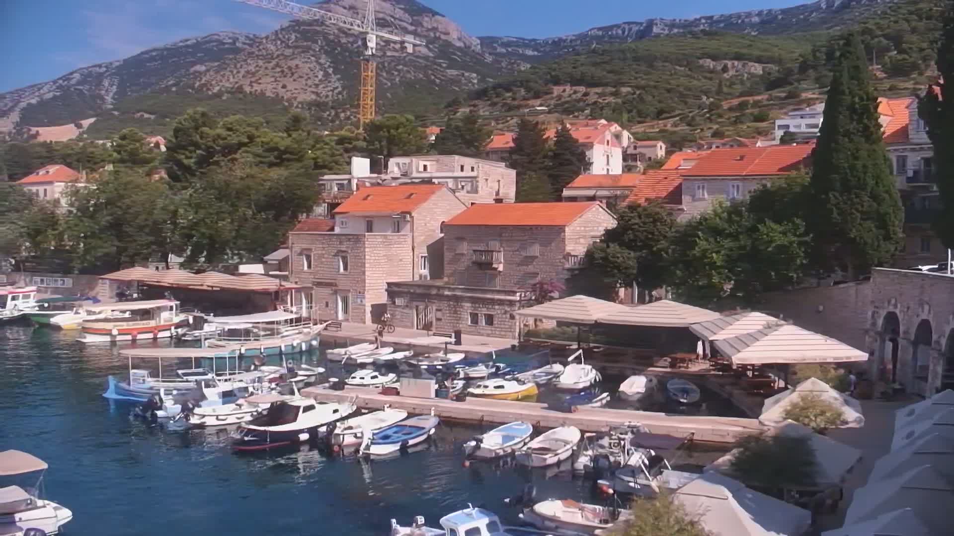 A harbor filled with numerous small boats is nestled in a village of stone buildings with red-tiled roofs, backed by lush green hills under a clear, sunny sky.