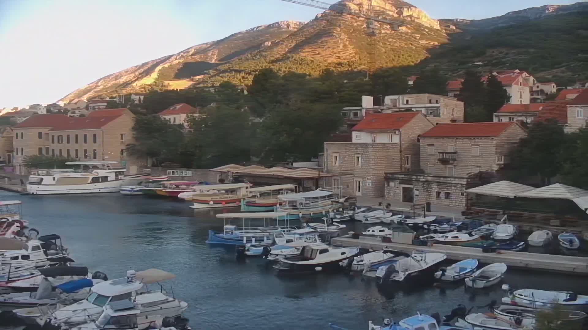 A harbor filled with numerous small boats is nestled in a village of stone buildings with red-tiled roofs, backed by lush green hills under a clear, sunny sky.