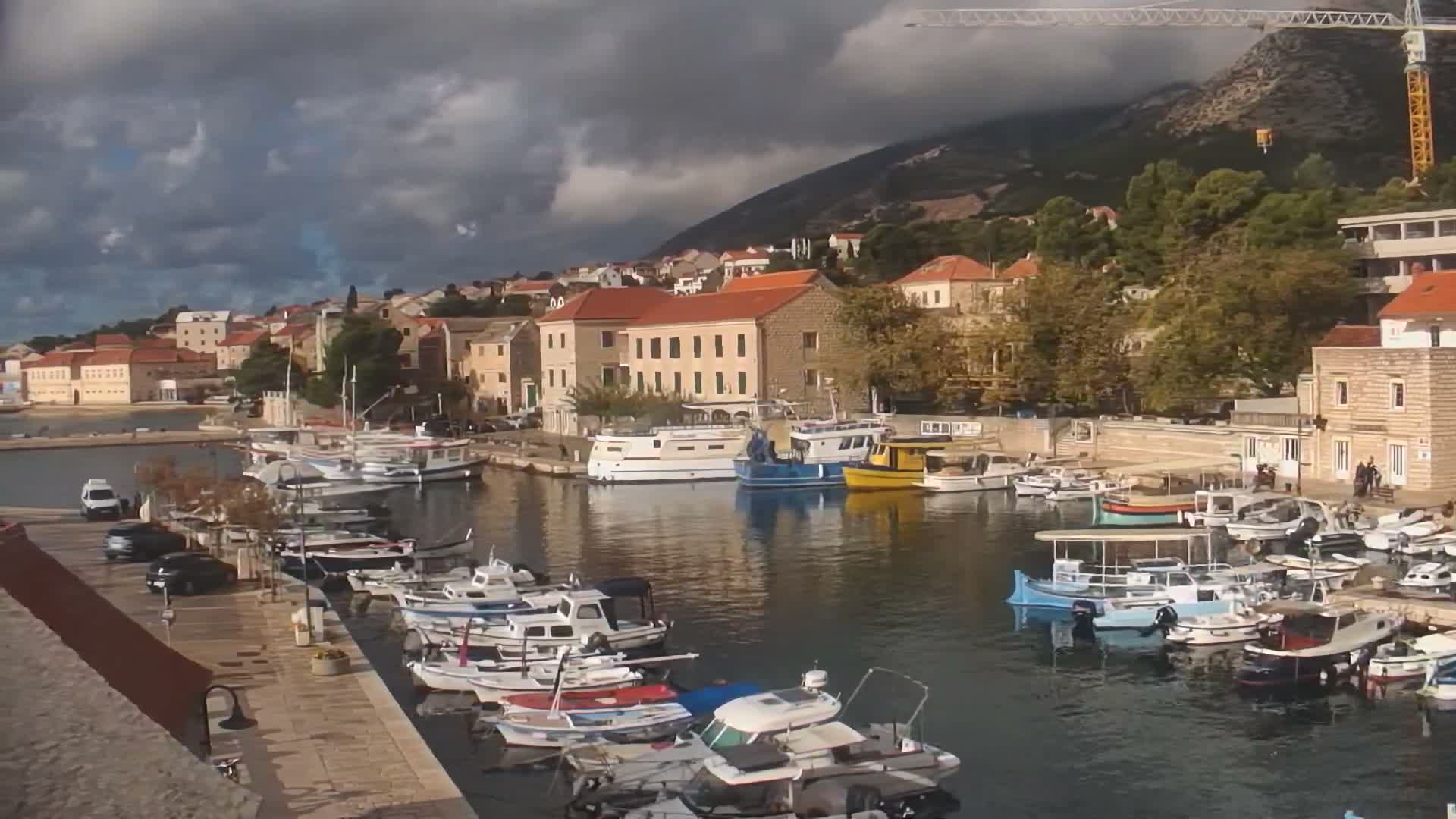 A picturesque harbor is filled with numerous boats docked along stone quays, fronting a charming town with terracotta roofs nestled against a steep mountainside under a dramatic, partly cloudy sky with patches of both sun and dark clouds.