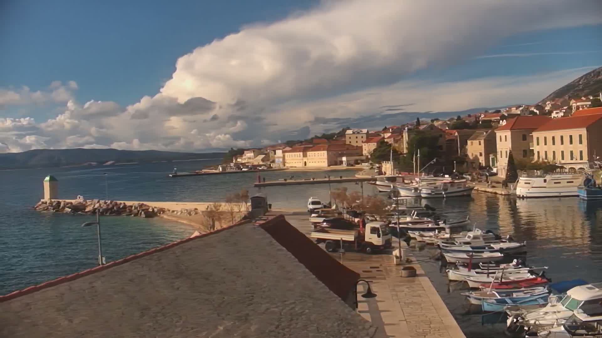 The image displays a picturesque coastal town with red-roofed buildings nestled against a hillside, a busy harbor filled with various boats, and a lighthouse on a rocky pier, all under a partly cloudy sky with bright sunshine.