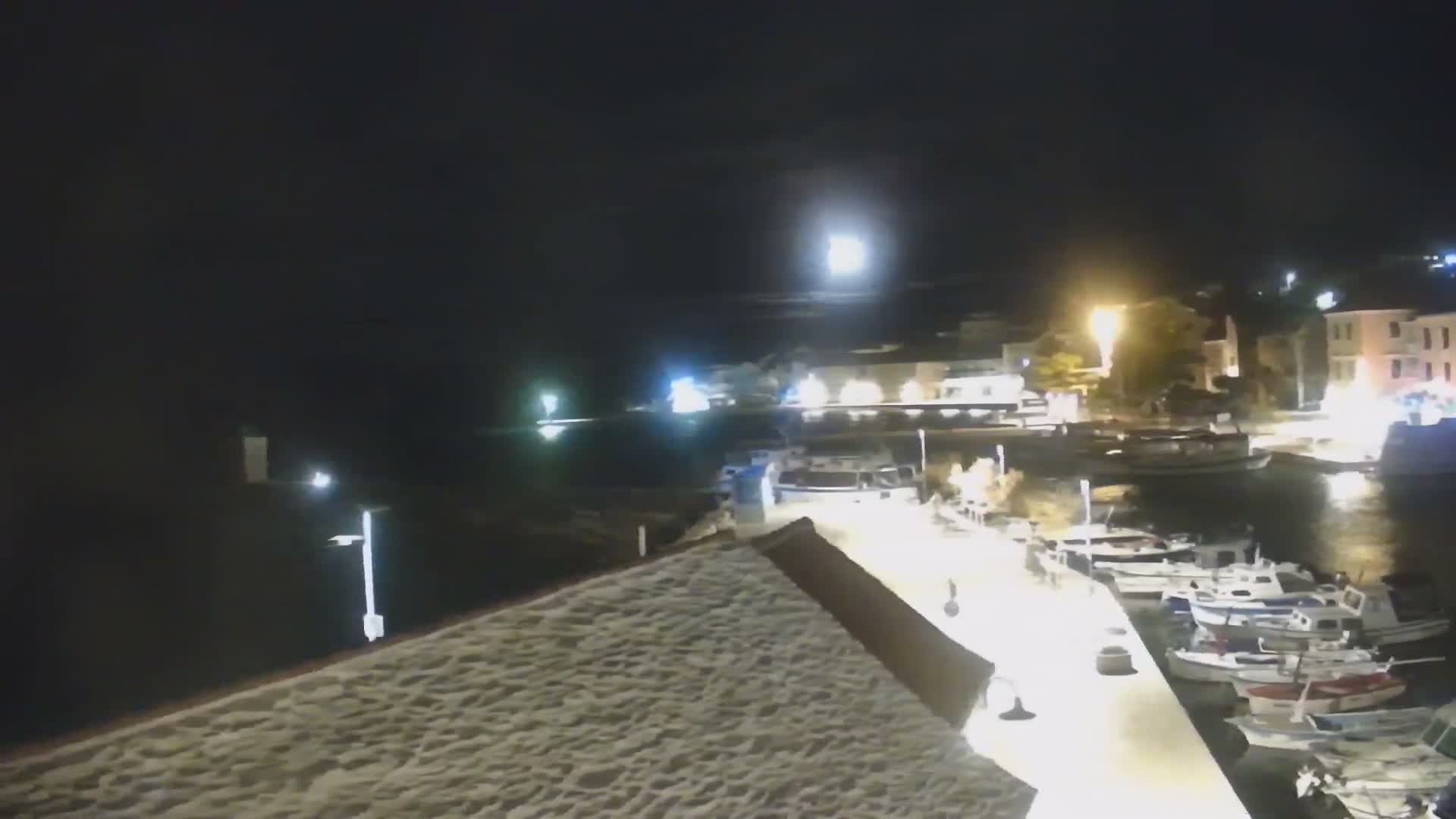 A clear night illuminates a harbor scene from an elevated view, featuring many boats docked along a brightly lit pier, with illuminated buildings visible across the water.