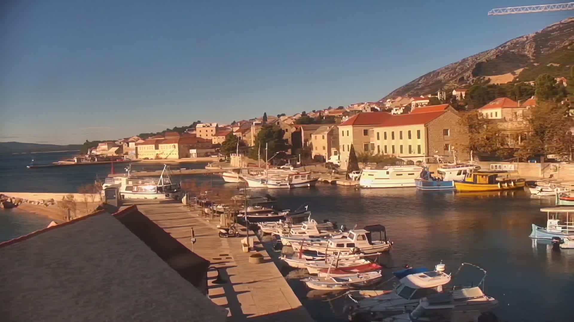 A picturesque coastal town with terracotta-roofed buildings climbs a steep, rocky hillside overlooking a calm harbor filled with numerous boats, all bathed in warm sunlight under a clear blue sky.