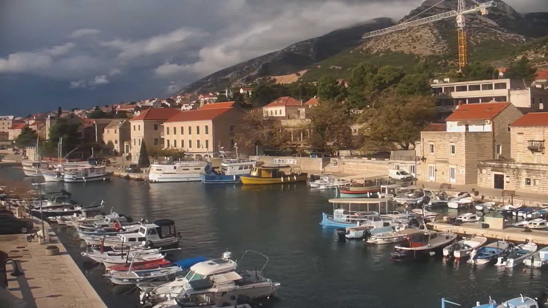 Numerous boats are moored in a scenic harbor bordered by a hillside town with red-roofed buildings and a large mountain featuring a prominent construction crane, all under a dramatic, partly cloudy sky.