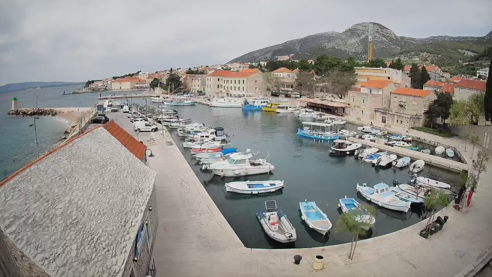 Numerous boats are moored in a scenic harbor bordered by a hillside town with red-roofed buildings and a large mountain featuring a prominent construction crane, all under a dramatic, partly cloudy sky.