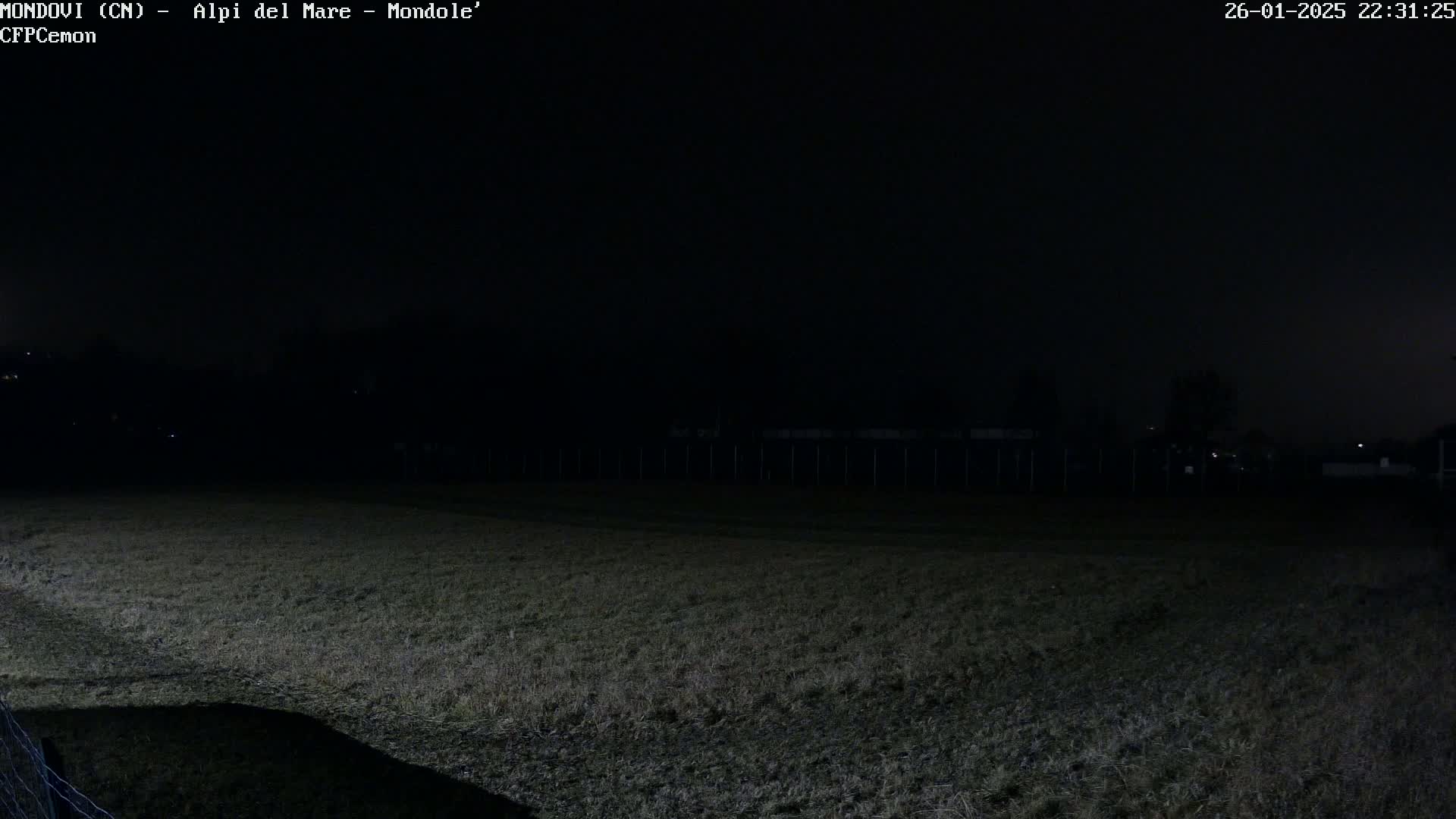 A dark, nighttime view of a grassy field, enclosed by a fence, under a mostly clear sky.