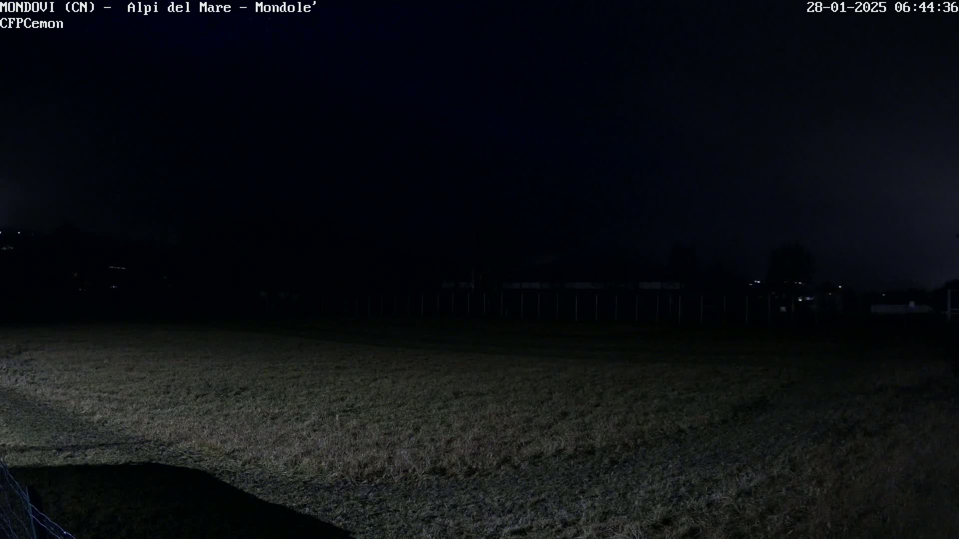 A dark, nighttime view of a grassy field with a fence and dimly lit structures in the background under a mostly clear sky.