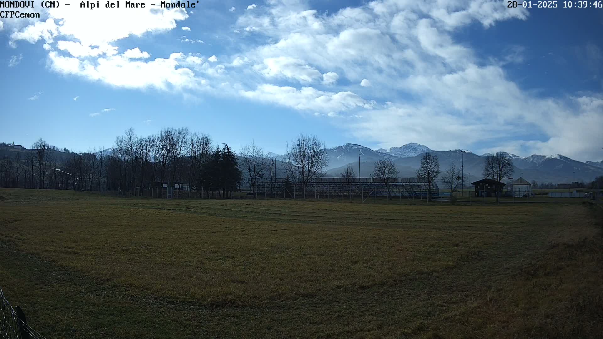 A grassy field with bleachers and bare trees sits in the foreground, with snow-capped mountains under a partly cloudy blue sky in the background.