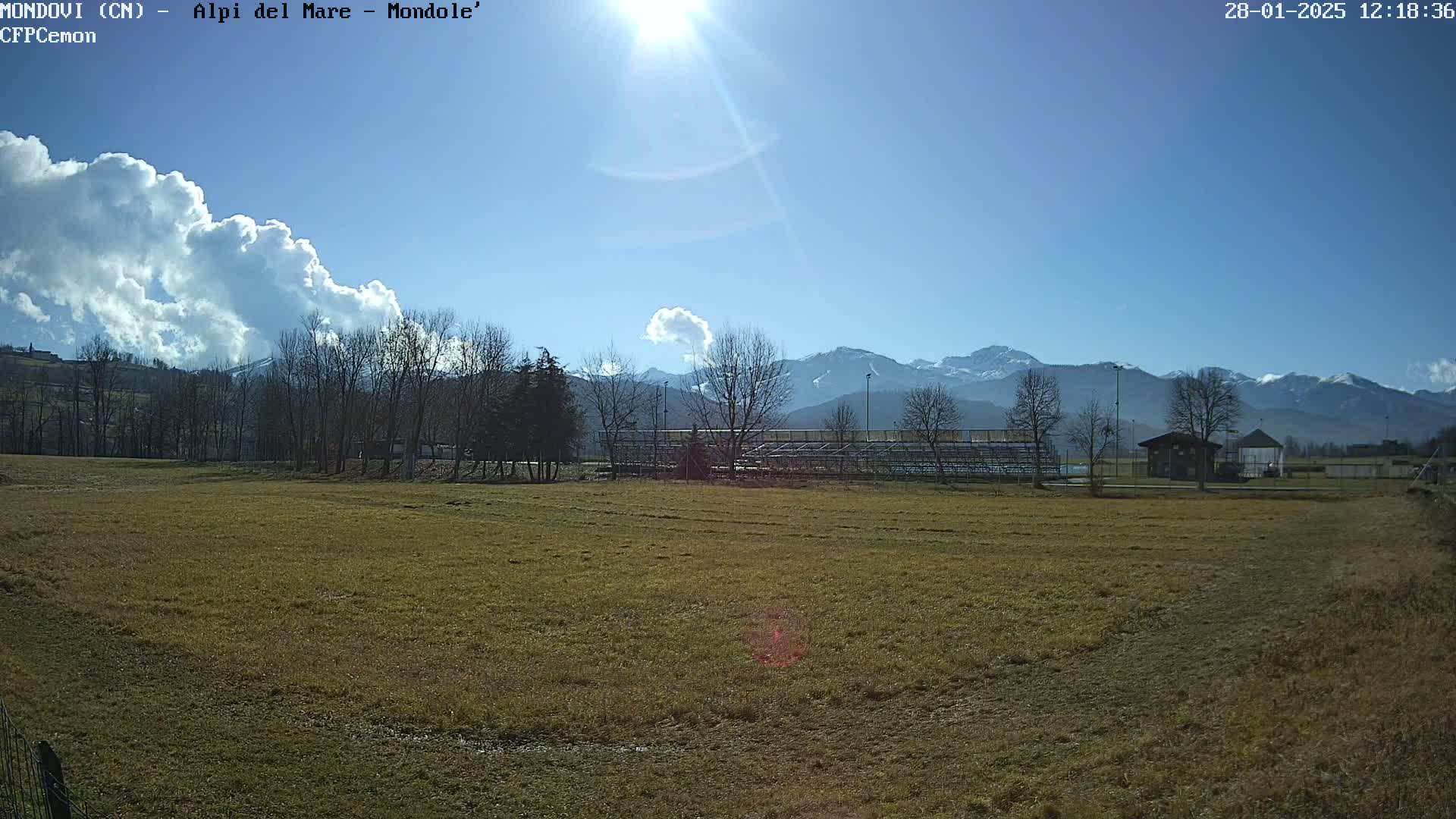 A sunny day reveals a grassy field, a partially covered bleacher structure, and snow-capped mountains in the distance, with a few fluffy clouds in the sky.