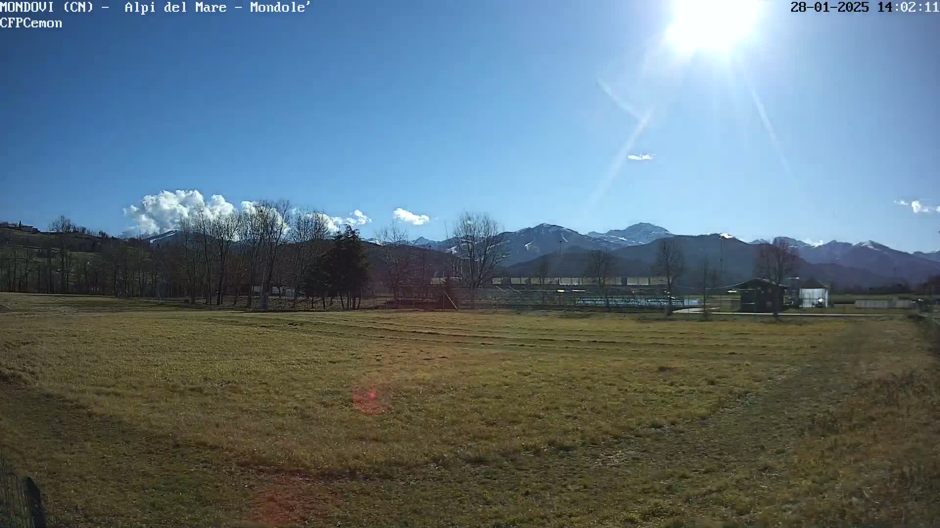 A sunny day reveals a brown field in the foreground, with leafless trees and distant snow-capped mountains under a mostly clear blue sky.