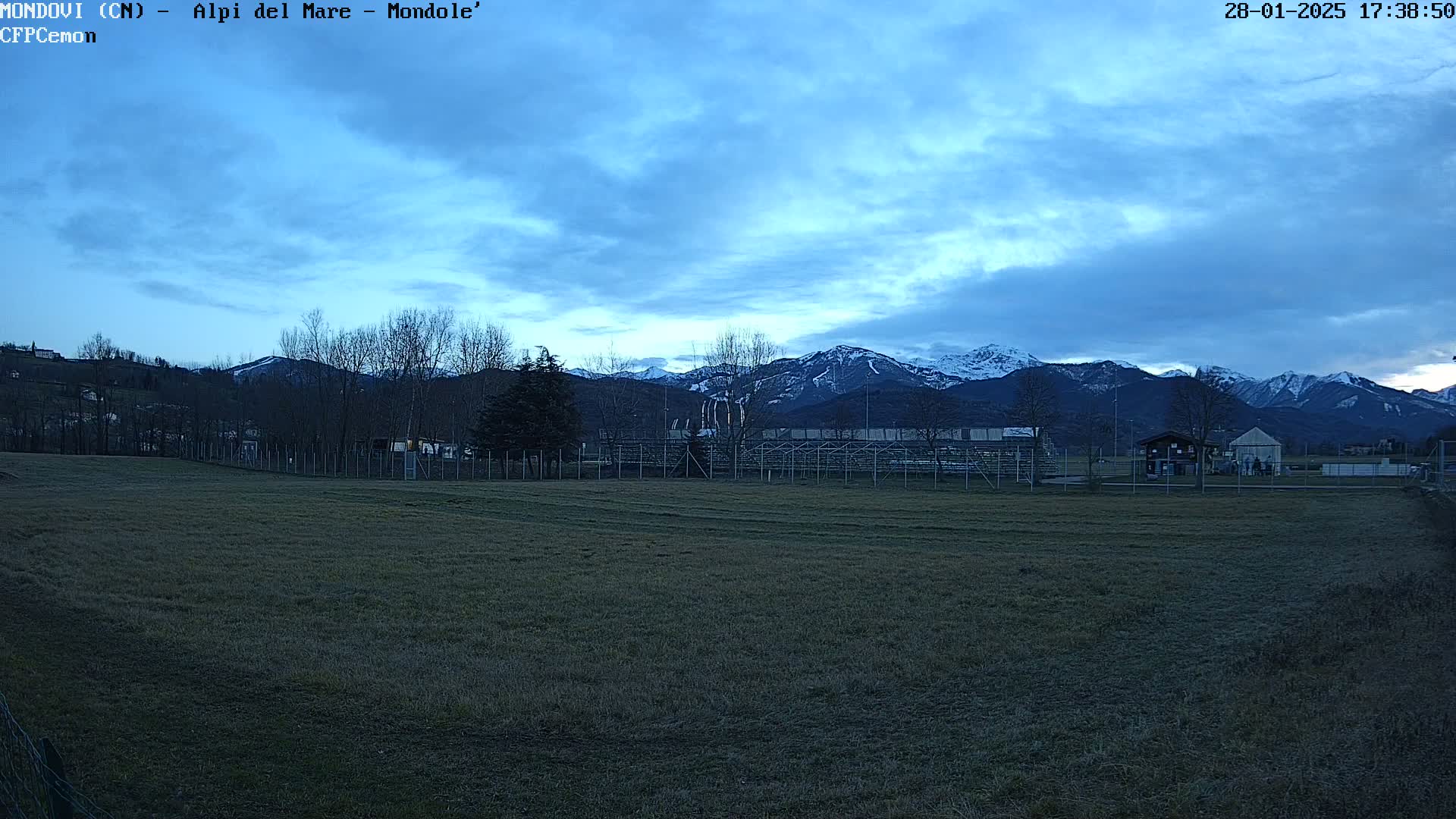 A mostly cloudy, twilight sky overlooks a grassy field with bleachers and a small building, all situated in front of snow-capped mountains.