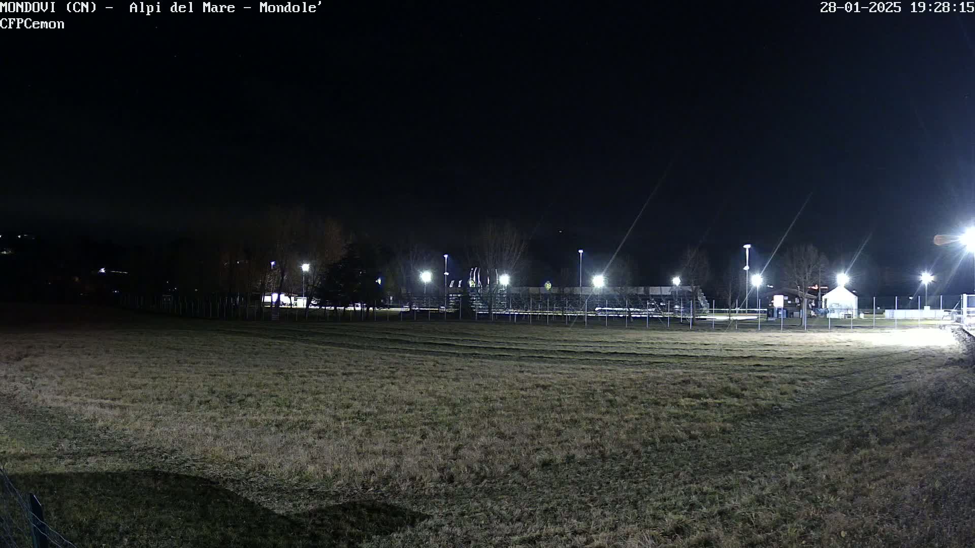 A nighttime view of a grassy field with a fenced-in area containing lights and structures in the background under a clear, dark sky.
