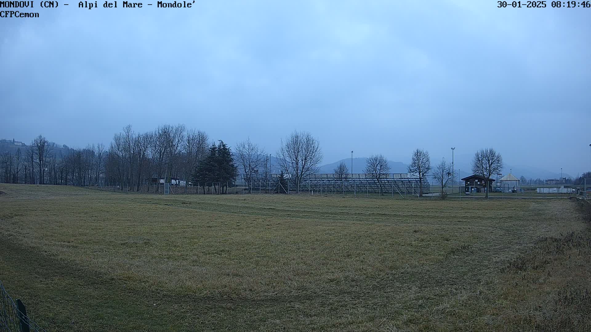 A field with short grass stretches towards bleachers and a small building under a cloudy sky.