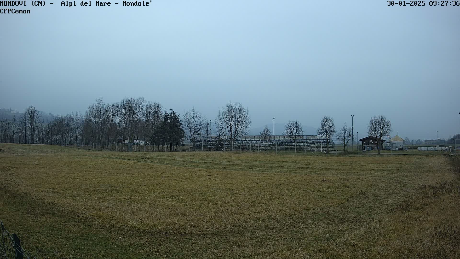 A brown grassy field stretches toward bleachers and bare trees under a hazy gray sky.