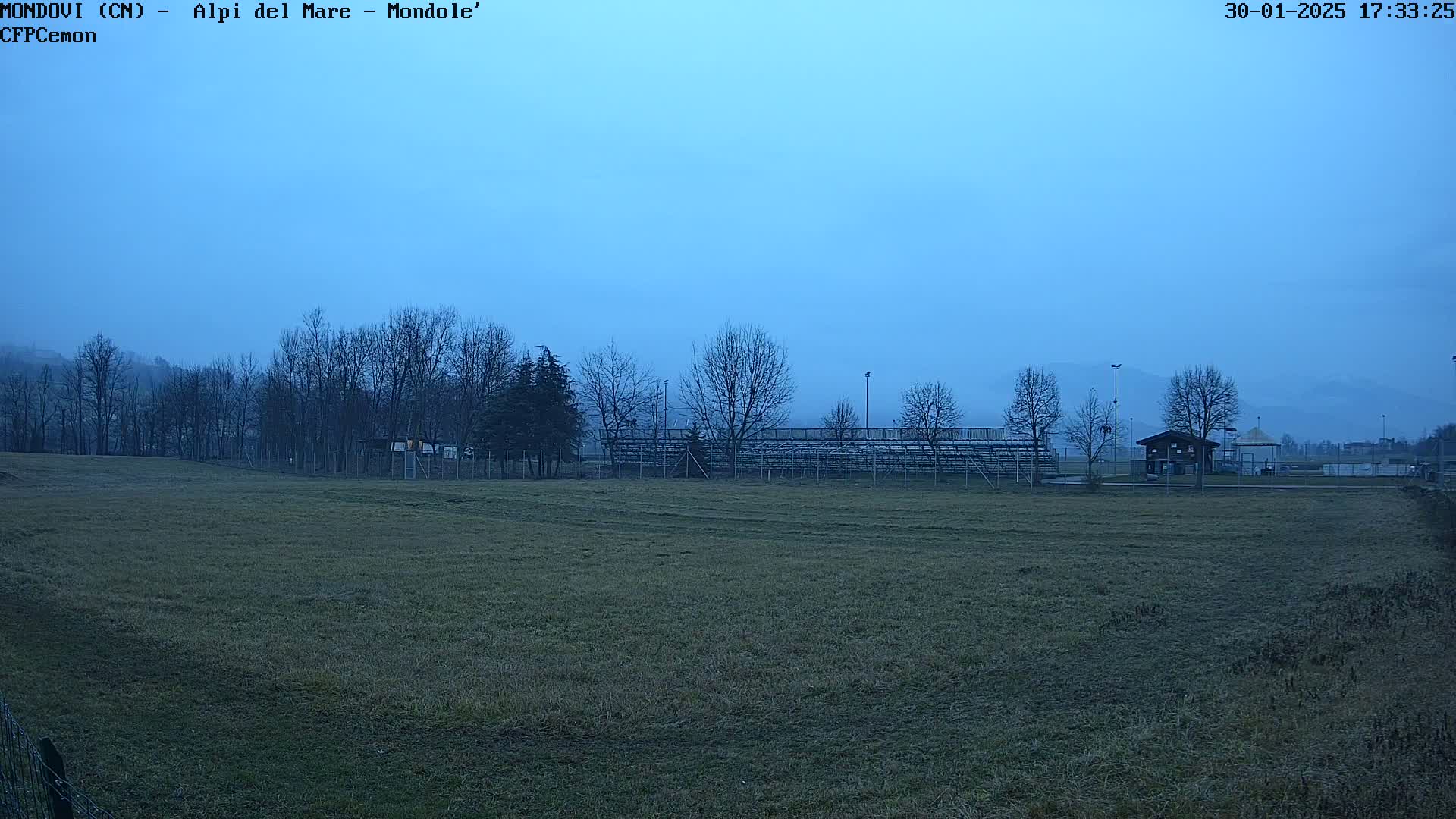 A mostly clear, dusky blue sky hangs over a grassy field with leafless trees, a small building, and empty bleachers in the background.