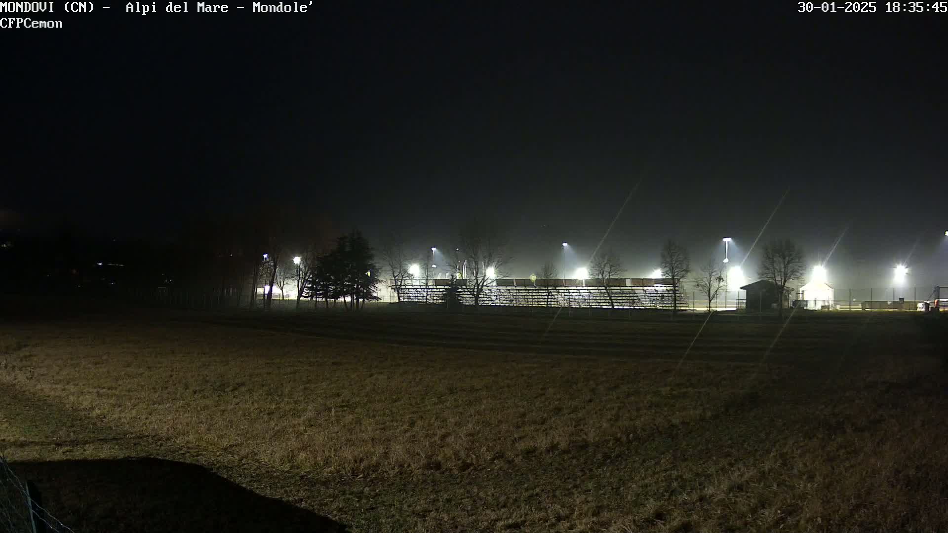 A nighttime, foggy view of a sparsely lit sports field with bleachers and a small building.