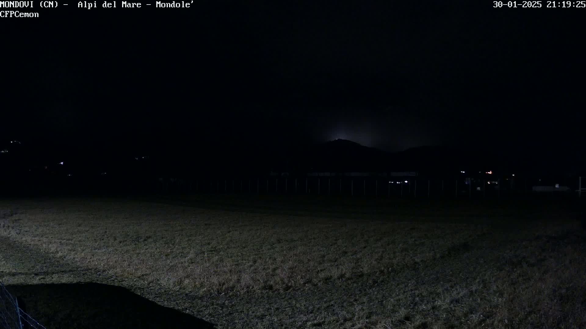 A dark, nighttime field stretches out before a dimly lit hill in the distance under a clear, moonlit sky.
