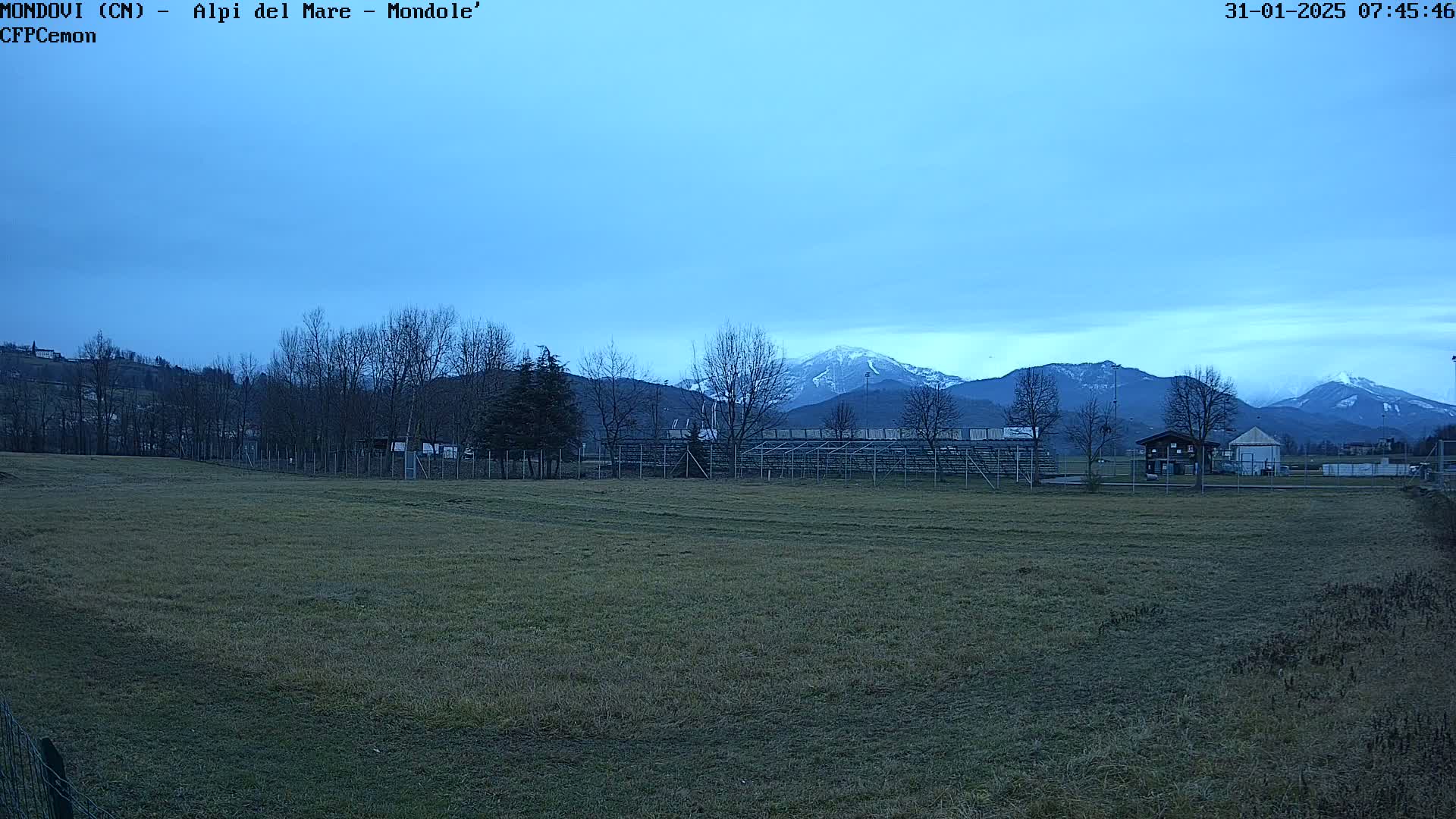 A grassy field with bleachers and a small building sits in the foreground, overlooking snow-capped mountains under a clear, twilight sky.