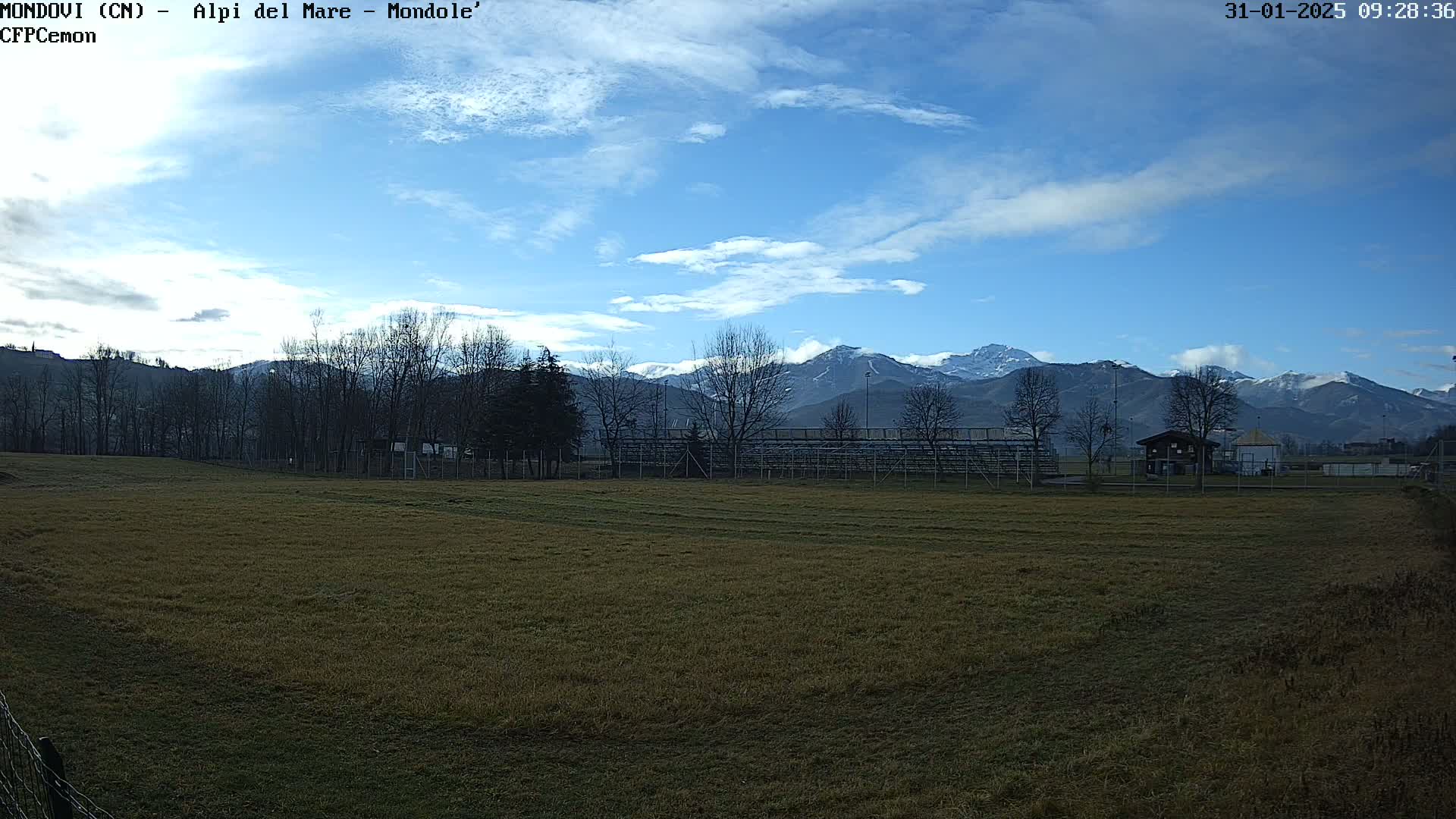 A mostly sunny day reveals snow-capped mountains in the distance behind a field with bleachers and bare trees.
