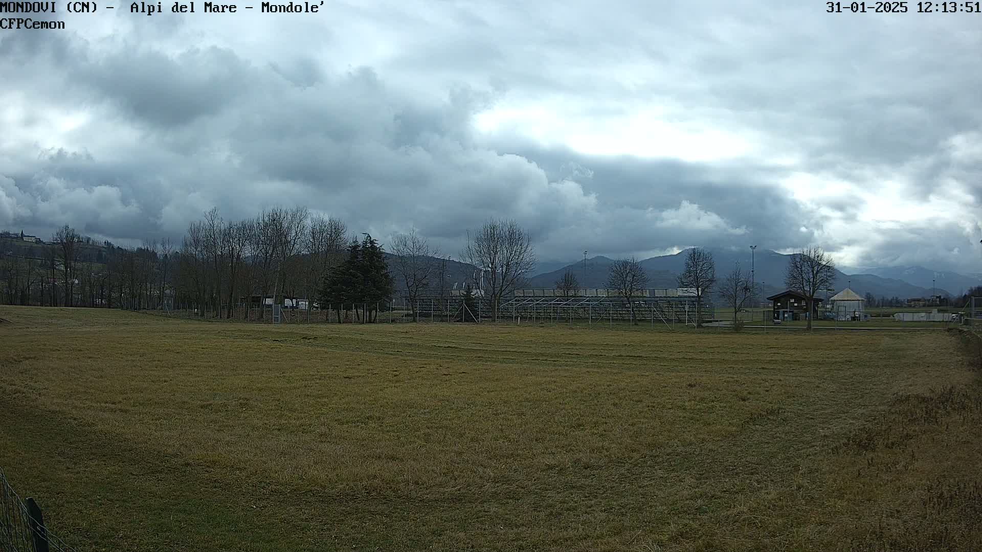 A mostly cloudy day reveals a brown field, bleachers, a few trees, and distant mountains.