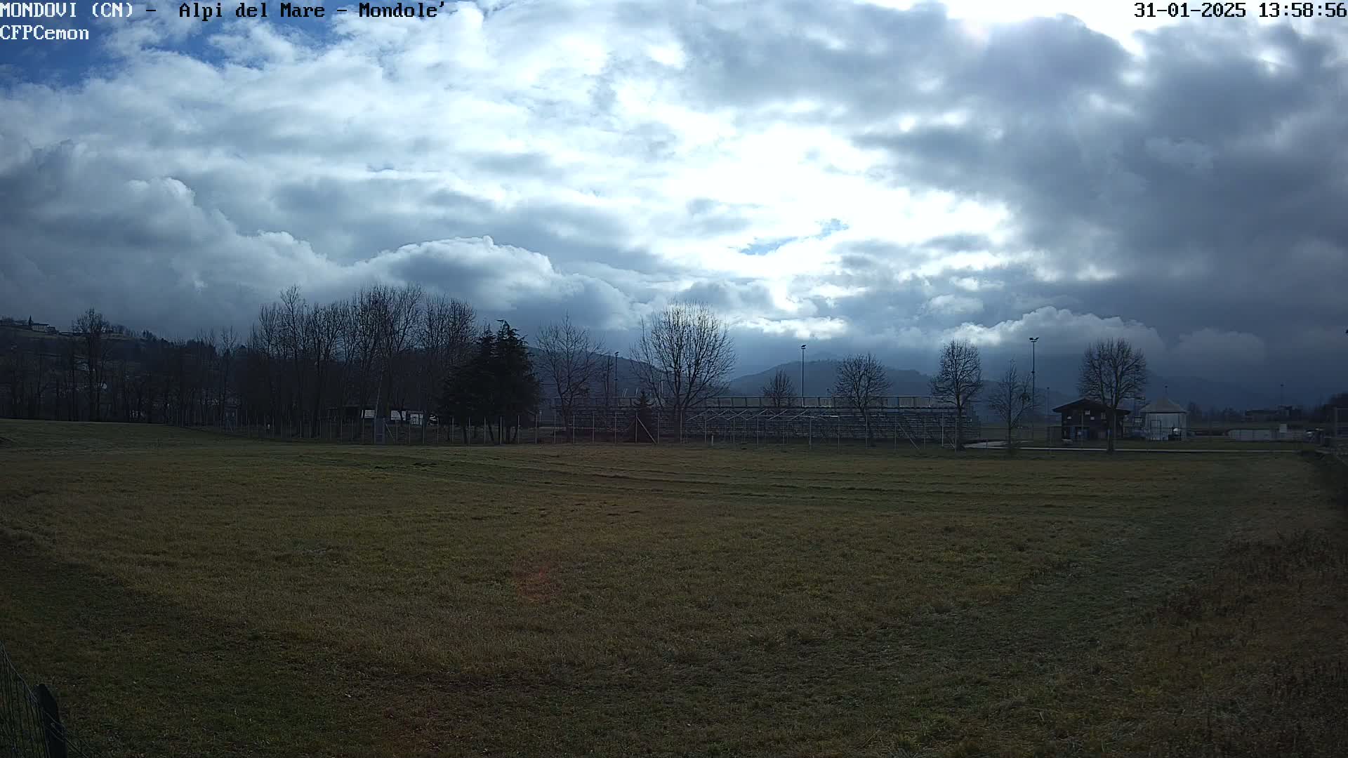 A mostly cloudy sky hangs over a field with bare trees and a small building, beyond which are distant mountains.