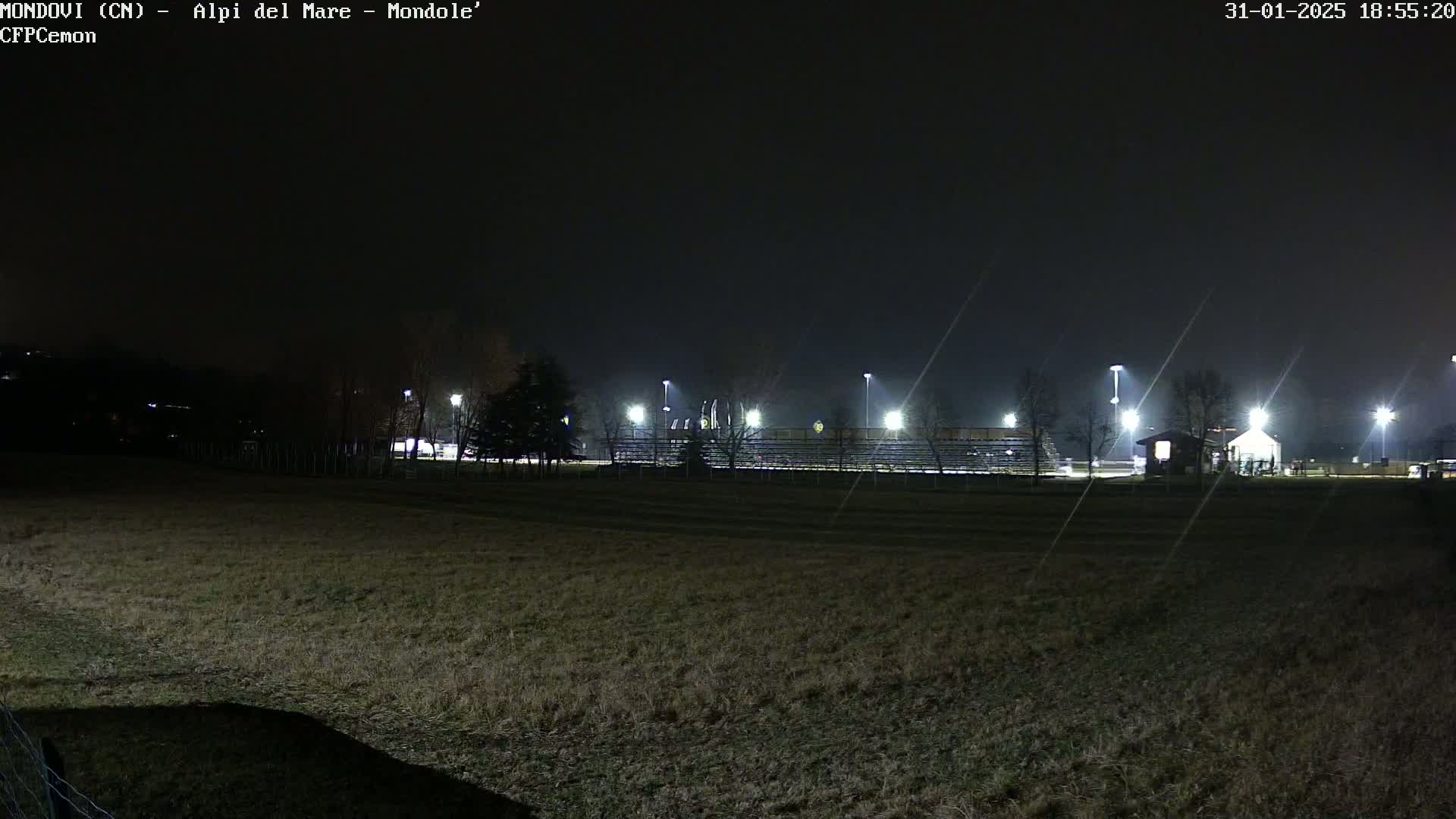 A nighttime view of an empty sports field with bleachers and floodlights under a dark, possibly foggy sky.