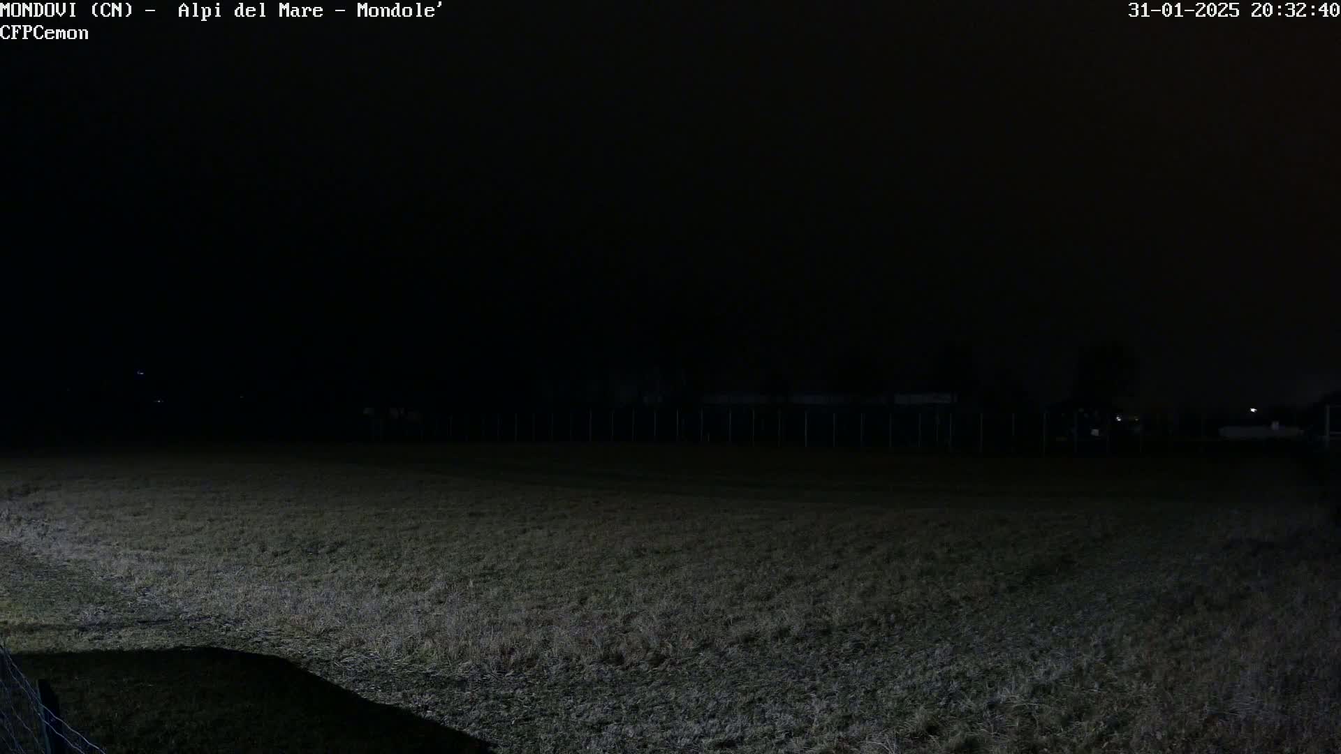 A dark, nighttime view of a grassy field, enclosed by a fence, with a few distant lights visible.